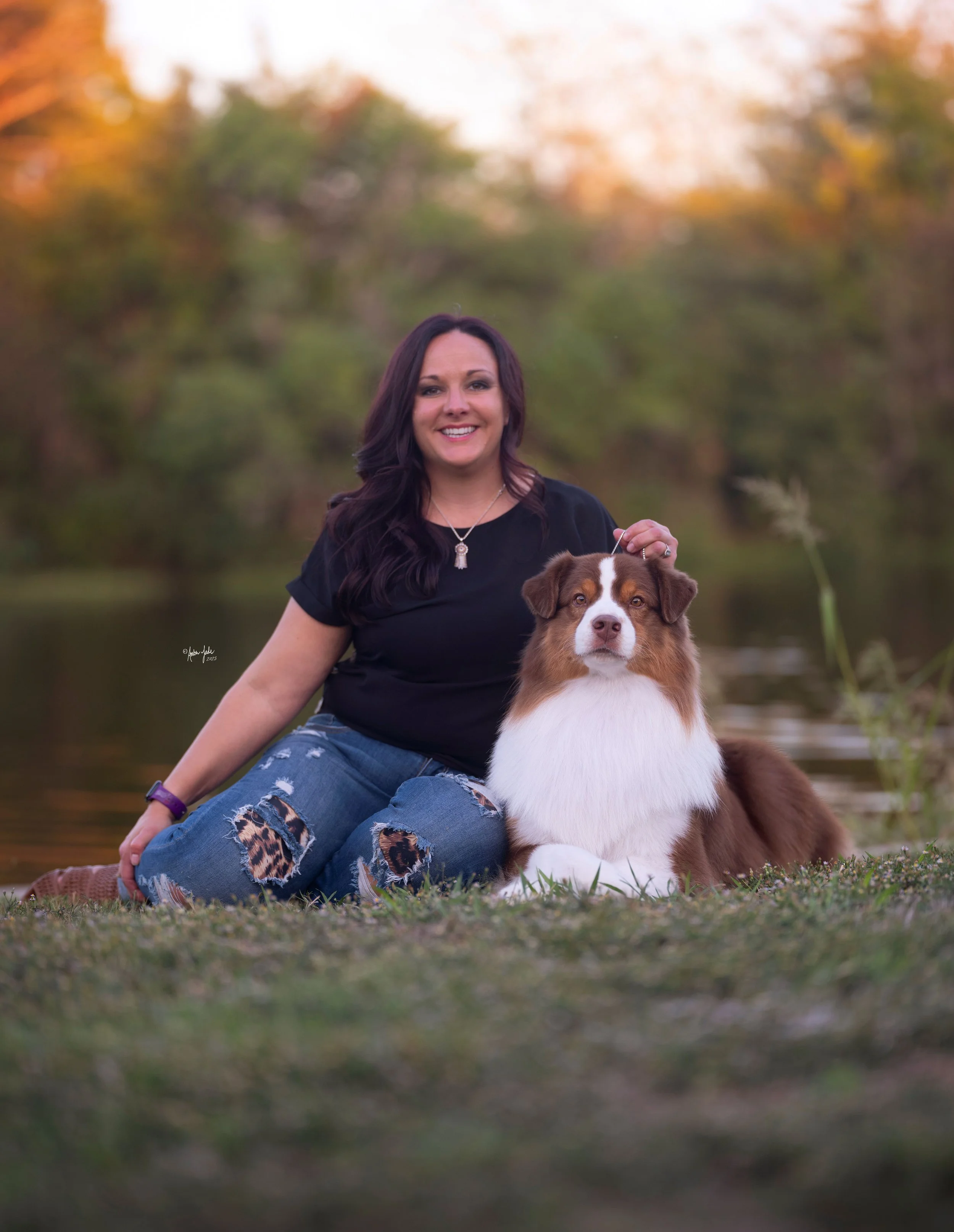 A woman with dark hair, wearing a black shirt and ripped jeans with leopard print patches, is sitting on the grass beside her red Australian Shepherd dog, near a body of water with a background of autumn trees.