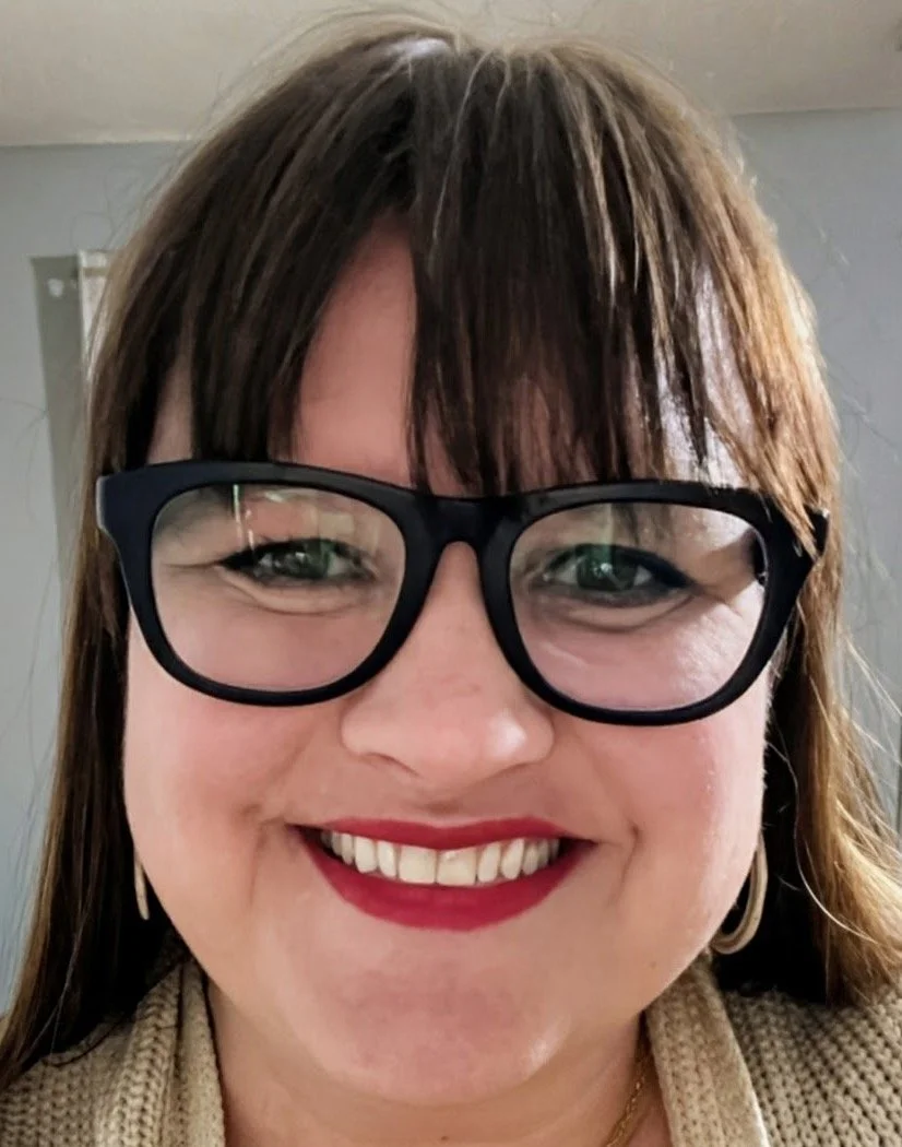 Tasha, a brunette wearing chunky black glasses, is smiling in a selfie.