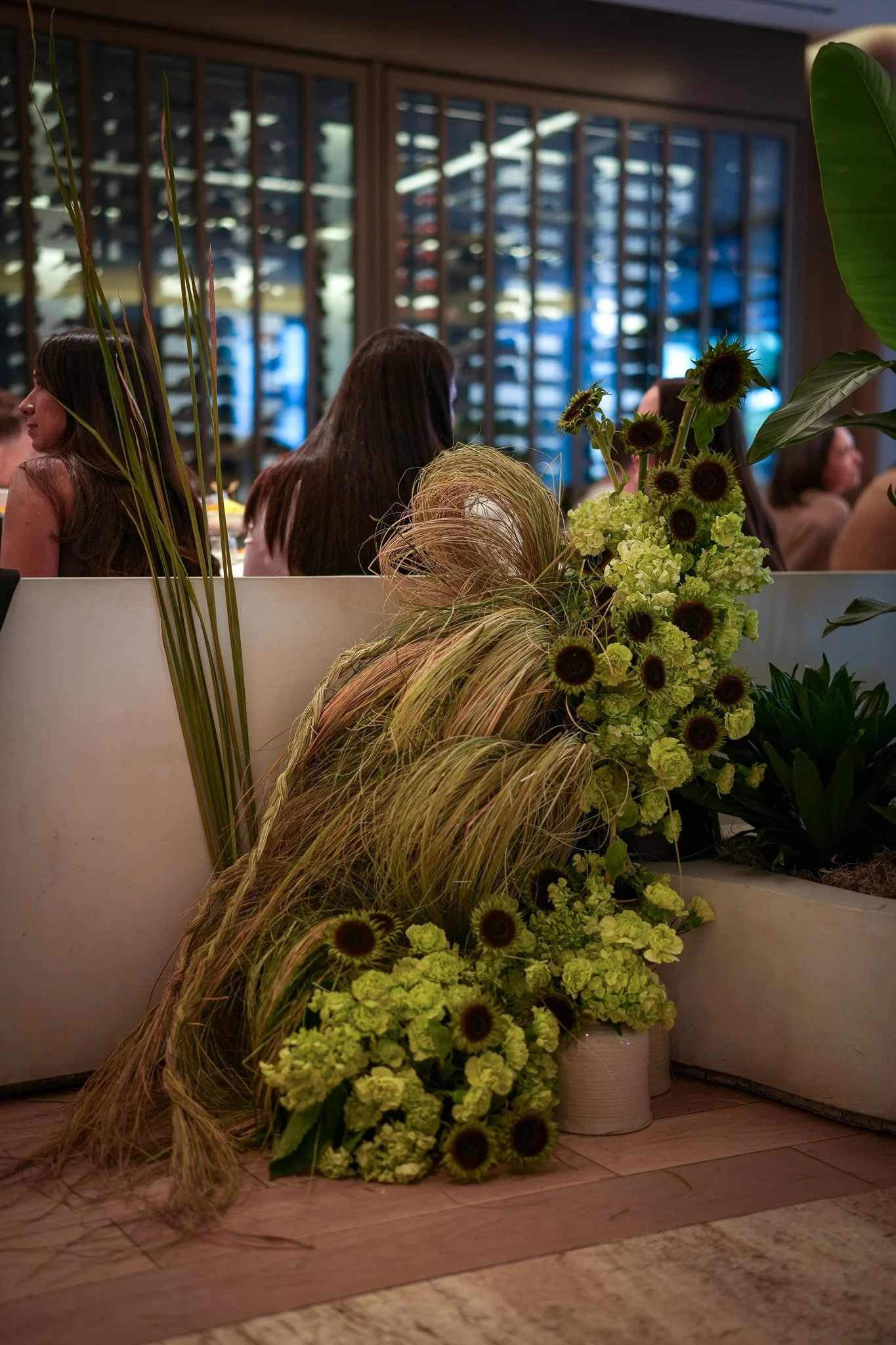 Un arrangement floral avec des tournesols, des hortensias vert clair, des graminées et des plantes vertes dans des vases blancs, placé près d'une barrière au sein d'un restaurant ou d'une salle de réception, avec des personnes assises en arrière-plan