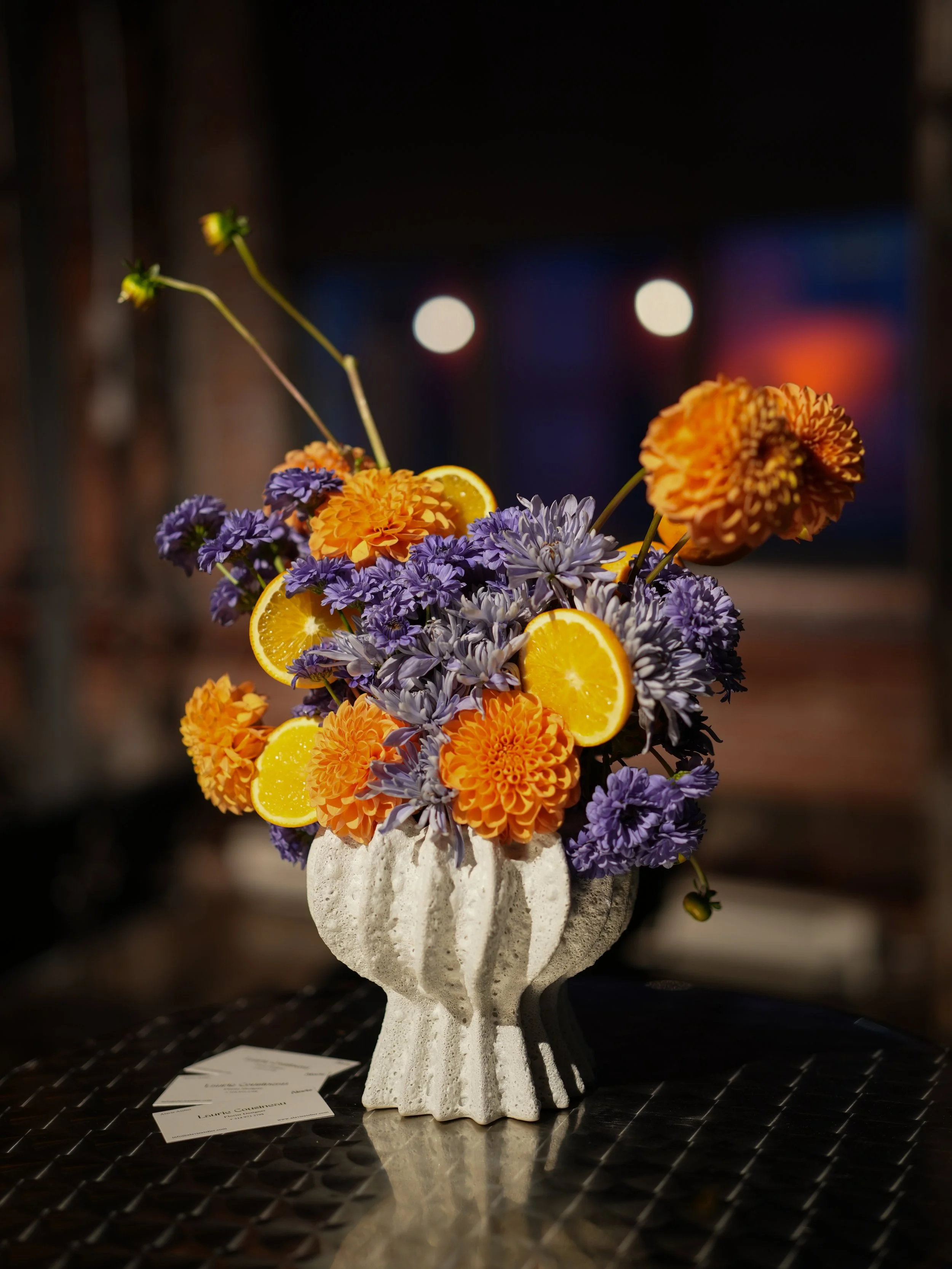 Fleurs colorées dans un vase en forme de main en pierre, avec des tranches d'orange, sur une table en métal, dans un environnement intérieur avec un éclairage chaud.