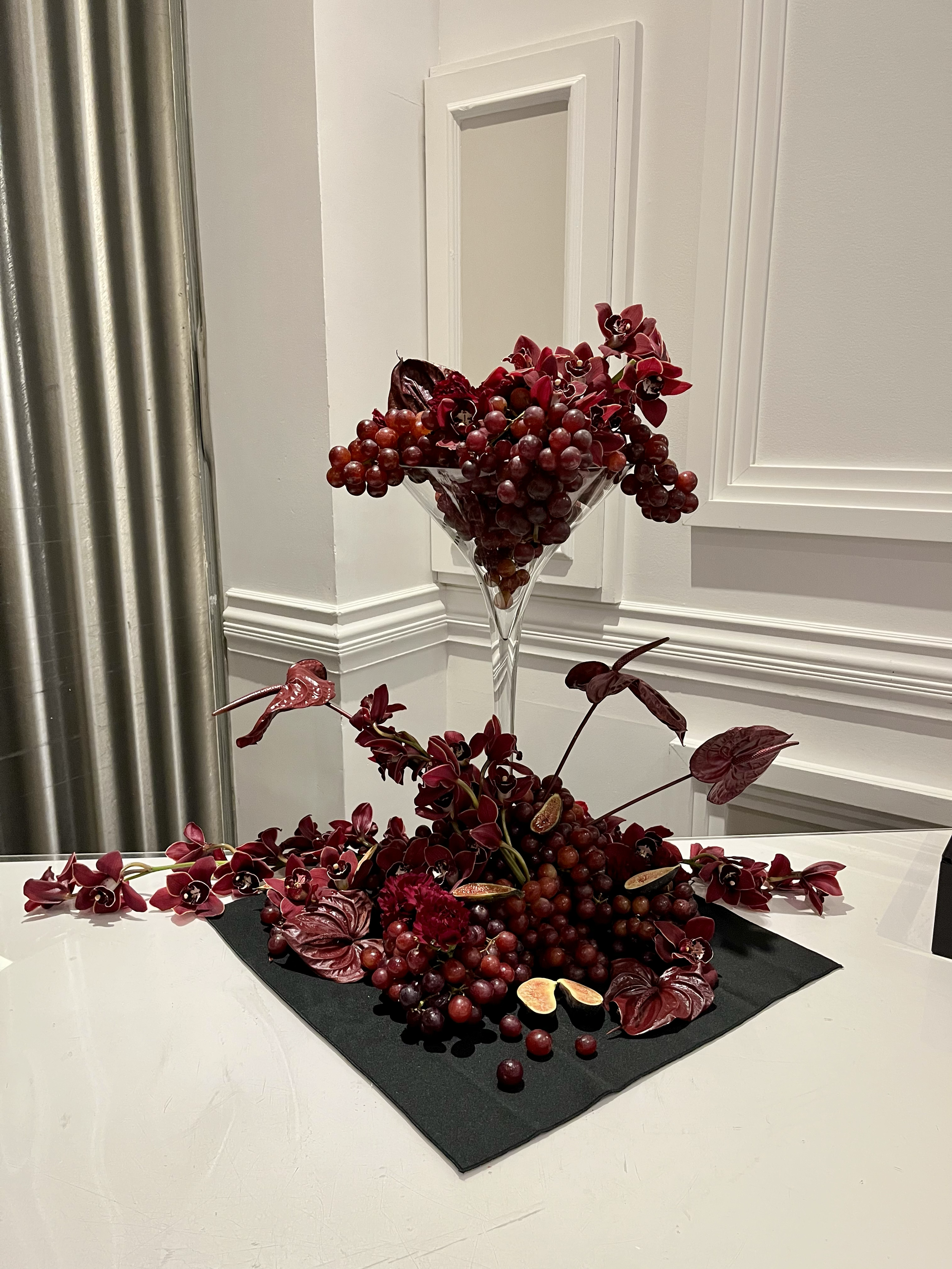 Une composition décorative de grappes de raisins rouges, de feuilles et de fleurs violettes dans un vase en verre placé sur une nappe noire blanche, en intérieur.