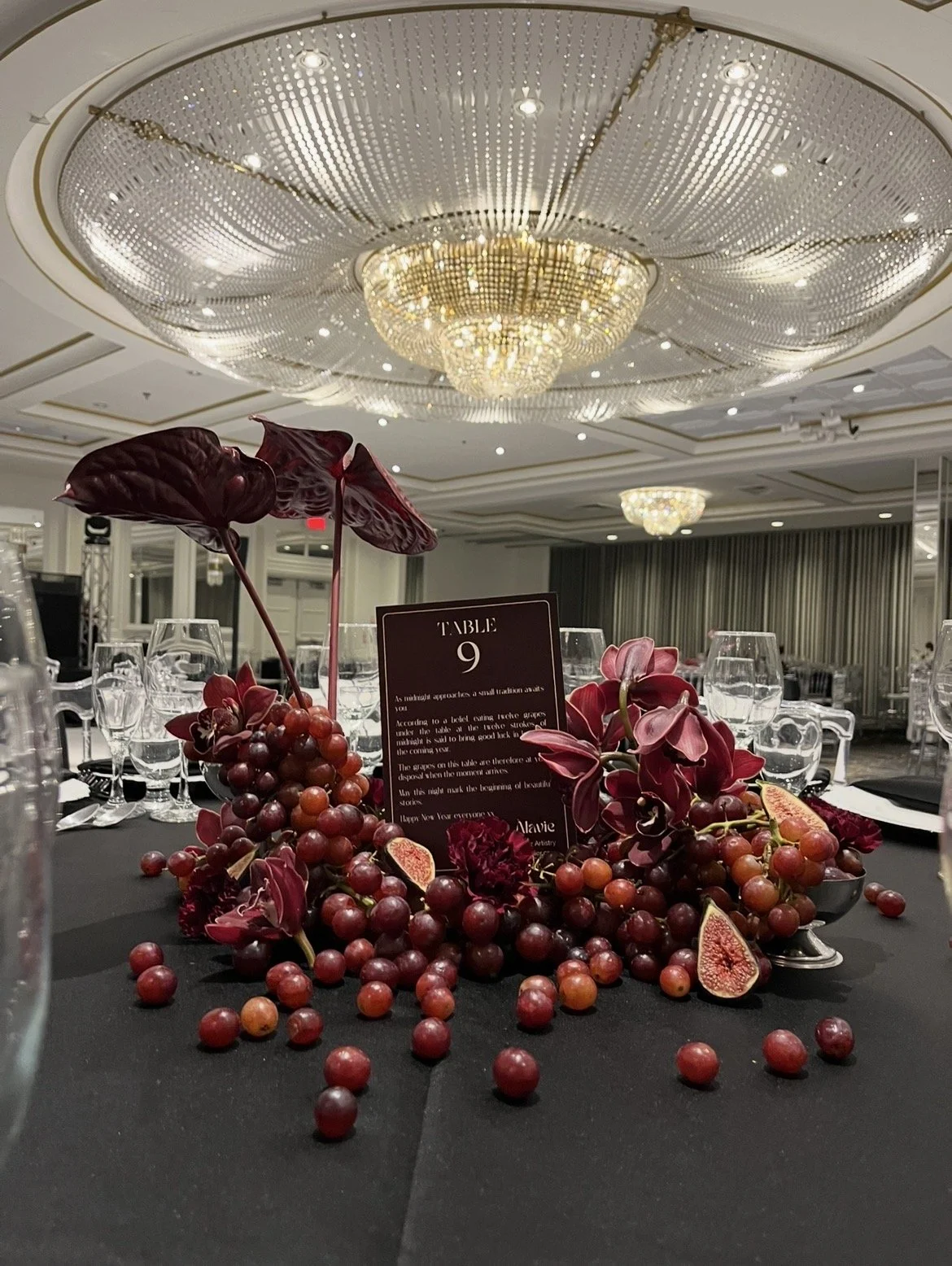 Décor de table élégante avec des grappes de raisins rouges, des fleurs pourpres, et un menu de table sur fond noir. L'ambiance est luxueuse, avec un plafond ornée de lustres en cristal dans une salle de réception.