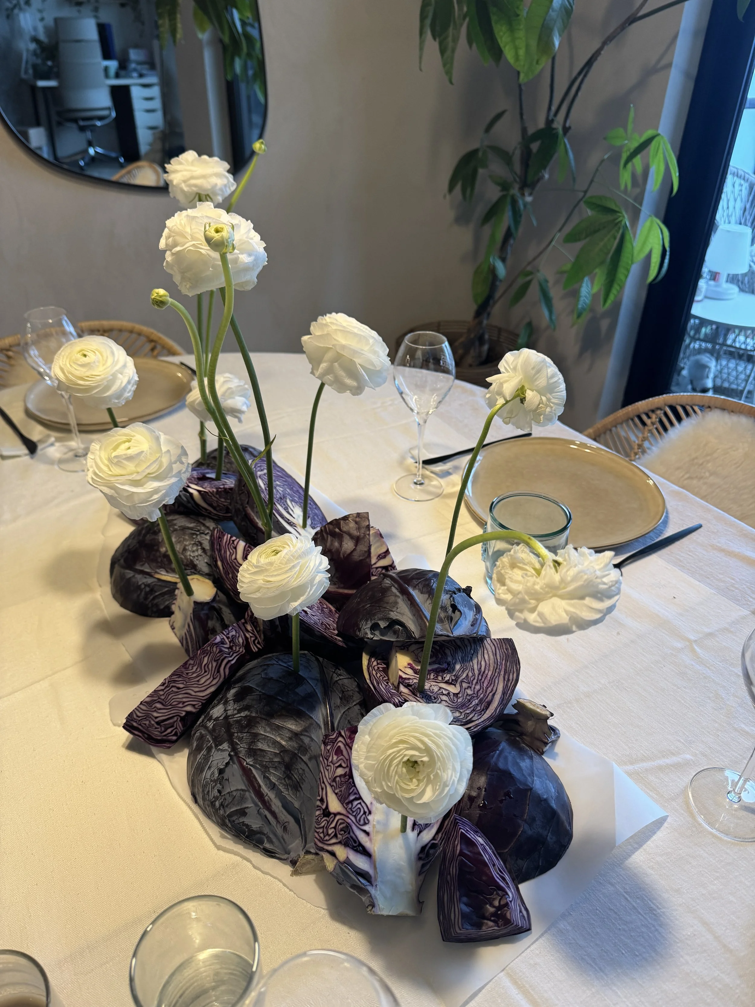 Centre de table avec des fleurs blanches et des morceaux de chou rouge sur une table avec des assiettes, des verres et des couverts dans une salle à manger.
