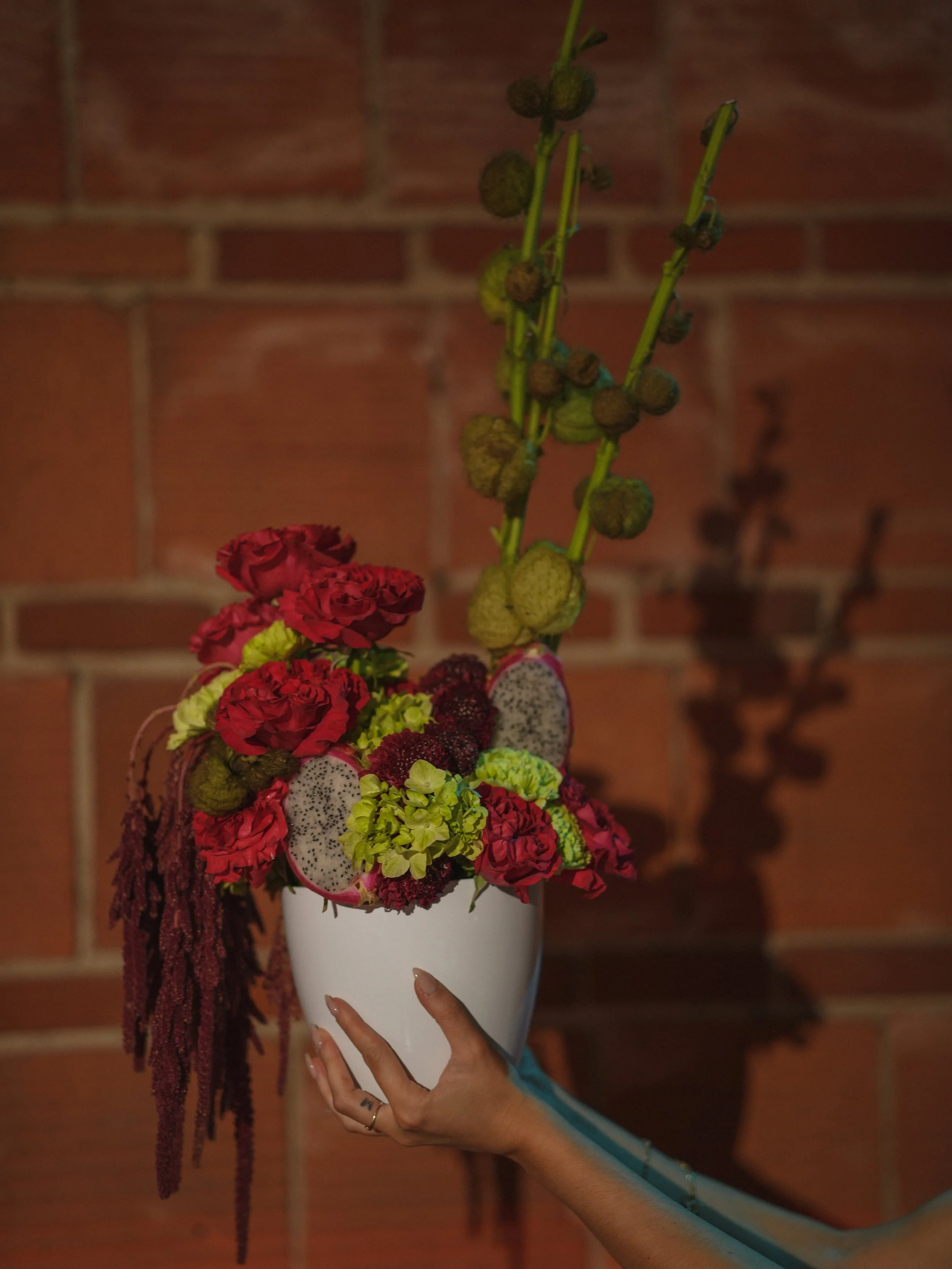 Main tenant un vase blanc avec un bouquet de fleurs rouges, vertes, et jaunes, comprenant des roses, des hortensias, des fougères et des fruits comme des grenades et du dragon fruit, contre un mur en briques