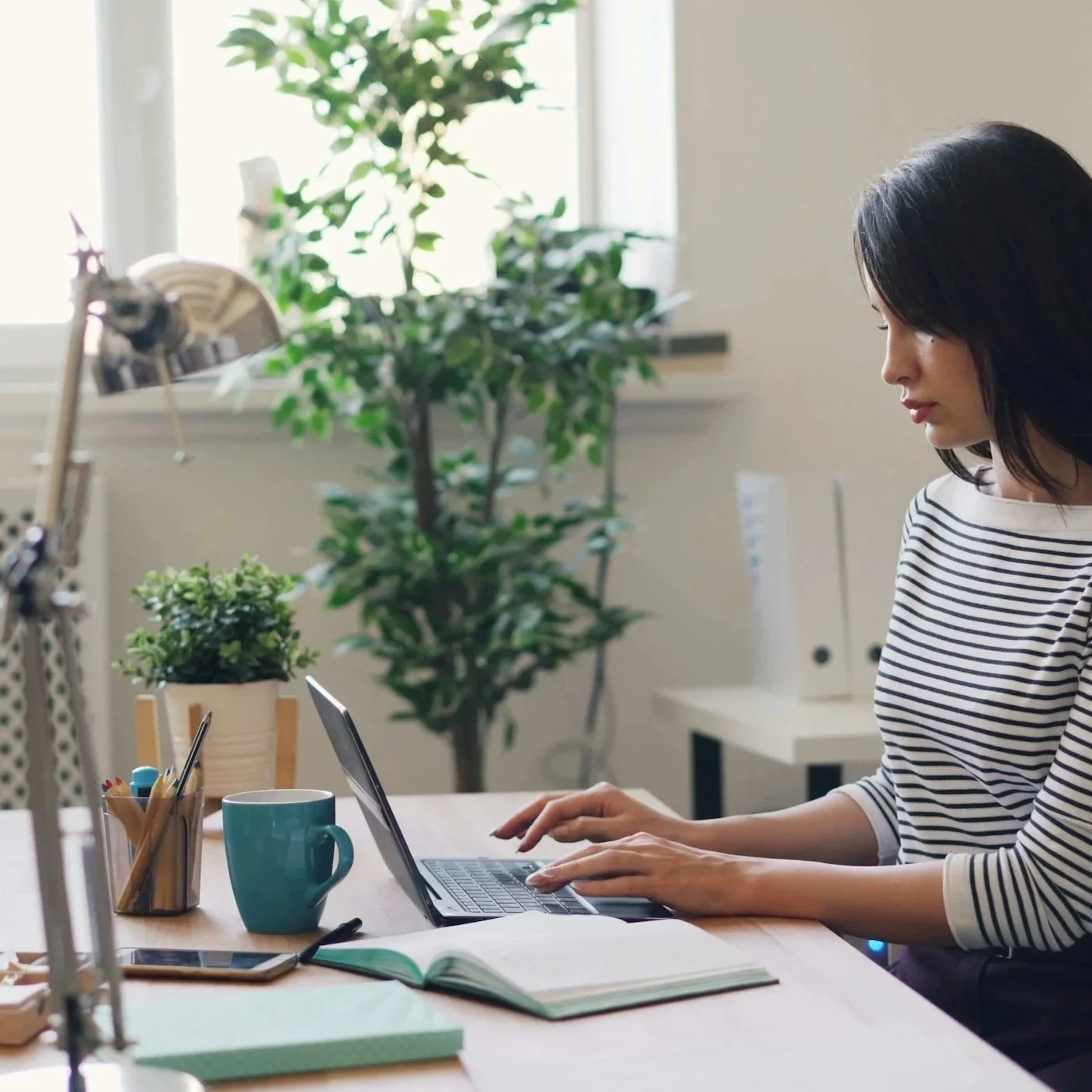 Woman working on a laptop at a desk in a bright room with a window, books, a mug, a potted plant, and office supplies.