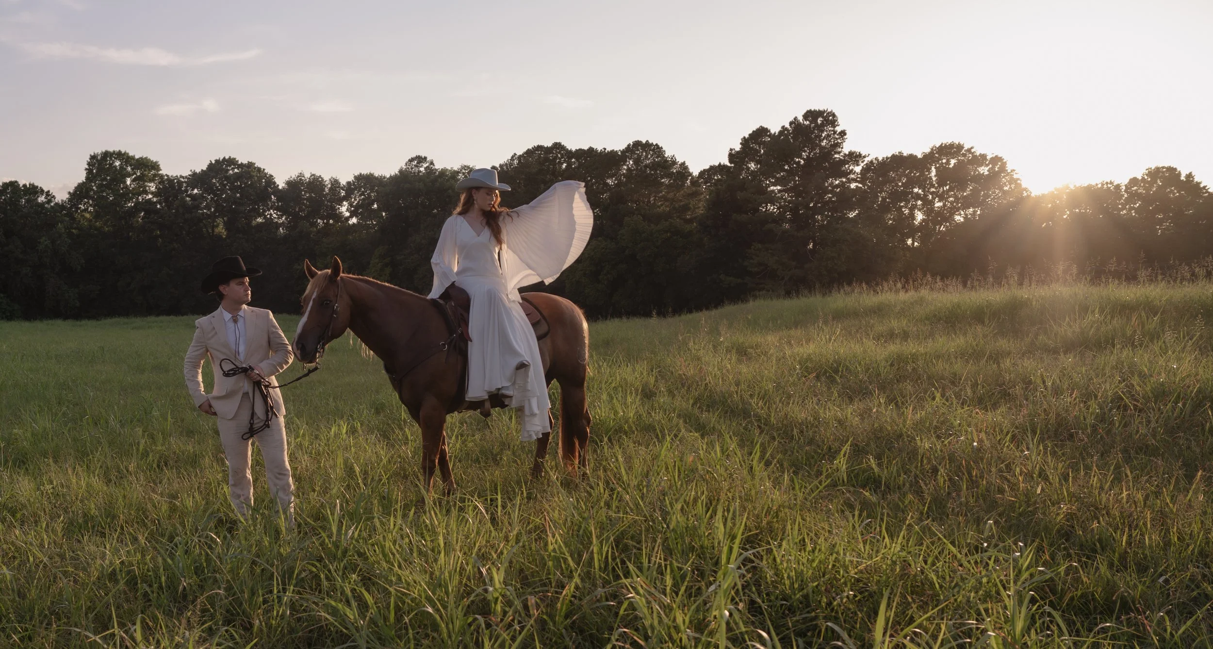 A woman in a white flowing dress and wide-brimmed hat sits on a brown horse in a grassy field at sunset, holding the reins being led by a man in a beige suit and black hat.