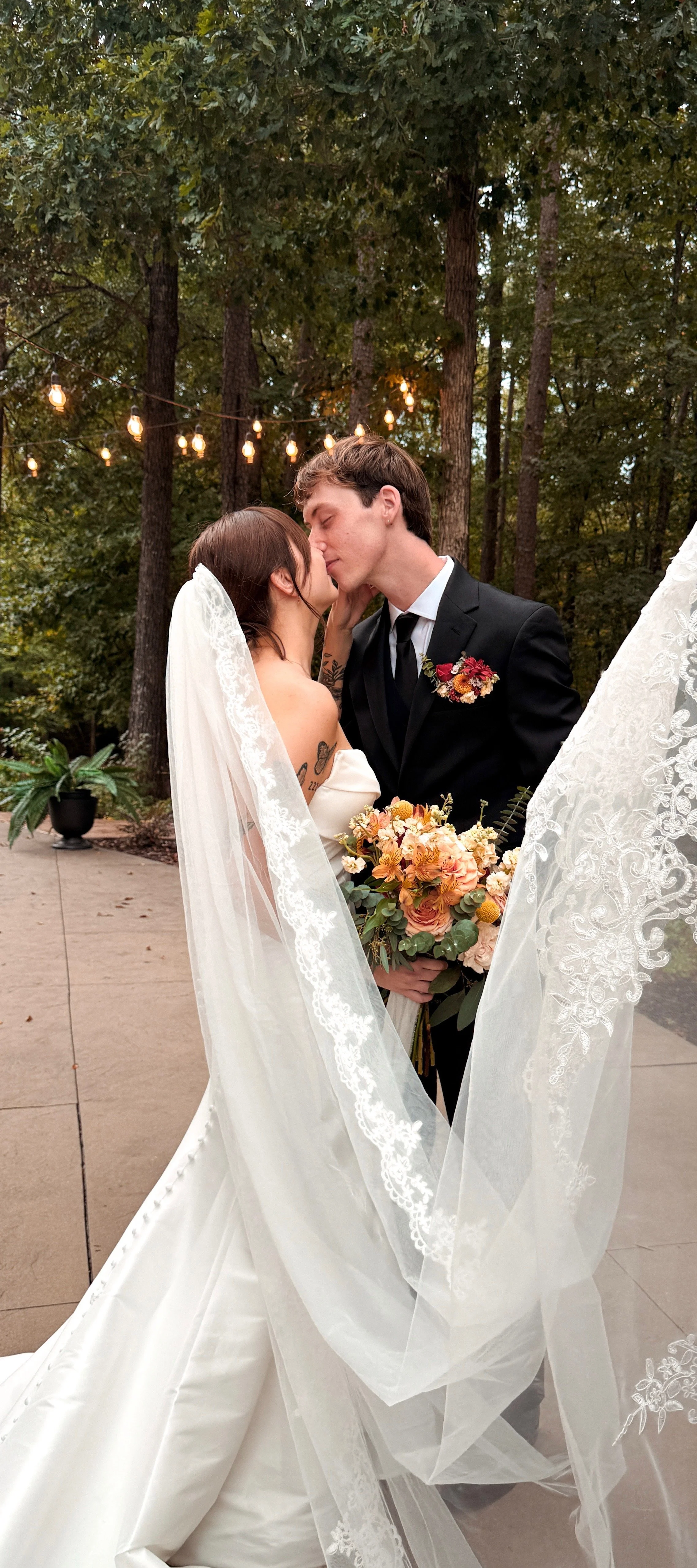 A bride and groom kiss during their outdoor wedding ceremony, surrounded by trees and string lights, with the bride holding a bouquet of flowers.
