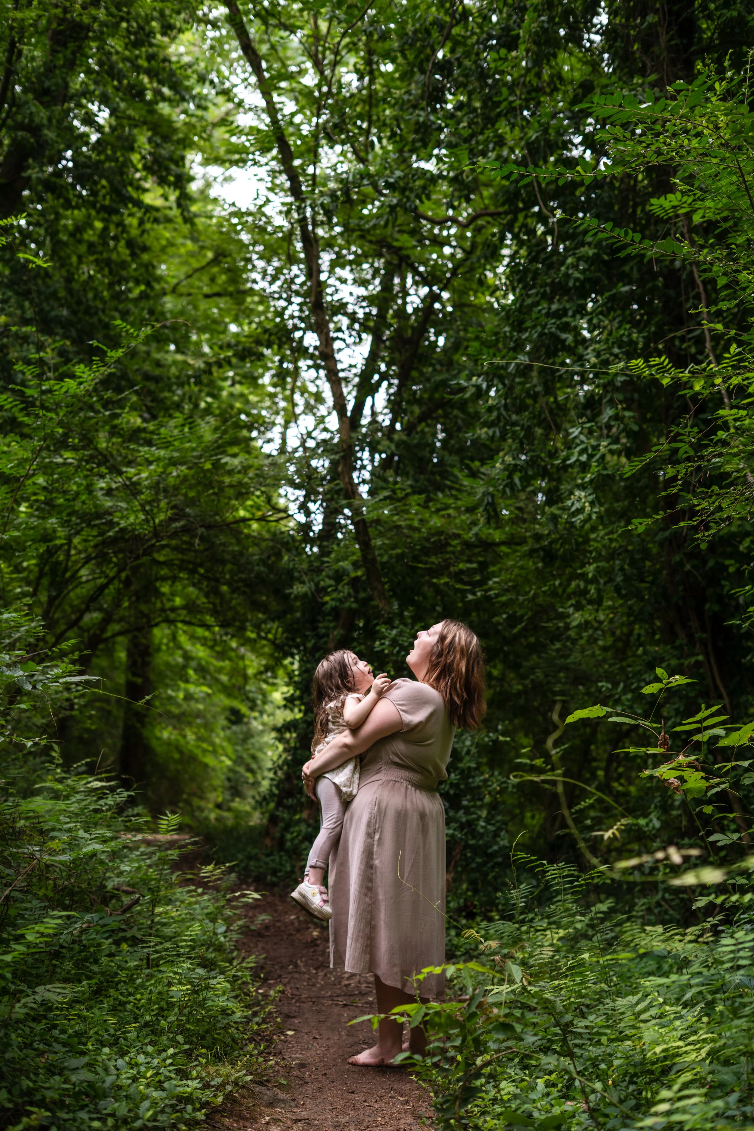 A woman holding a young girl in a forest with green trees, looking upward and smiling.