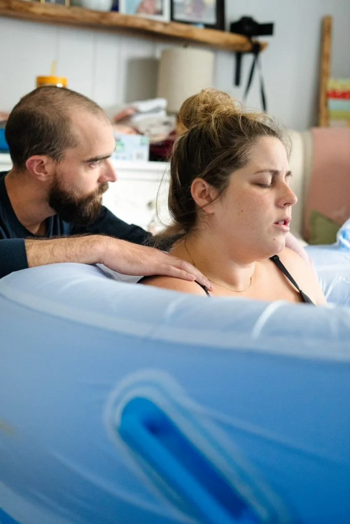 A woman appears to be in labor, with her eyes closed and an expression of discomfort or concentration. A man, possibly her partner, is beside her, touching her shoulder in support. They are in a room that resembles a home setting, with supplies and a shelf visible in the background.