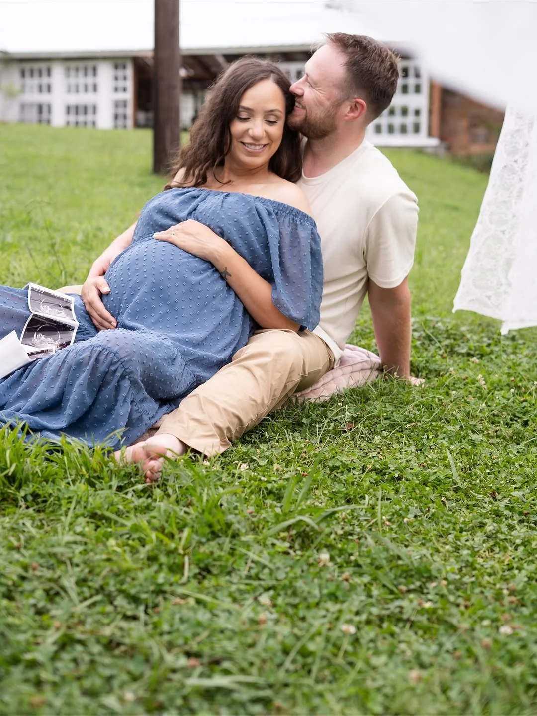 A pregnant woman in a blue dress sitting on grass, holding her belly, with a man next to her. They are outdoors, smiling, with ultrasound images near her. A white lace curtain is visible on the right.