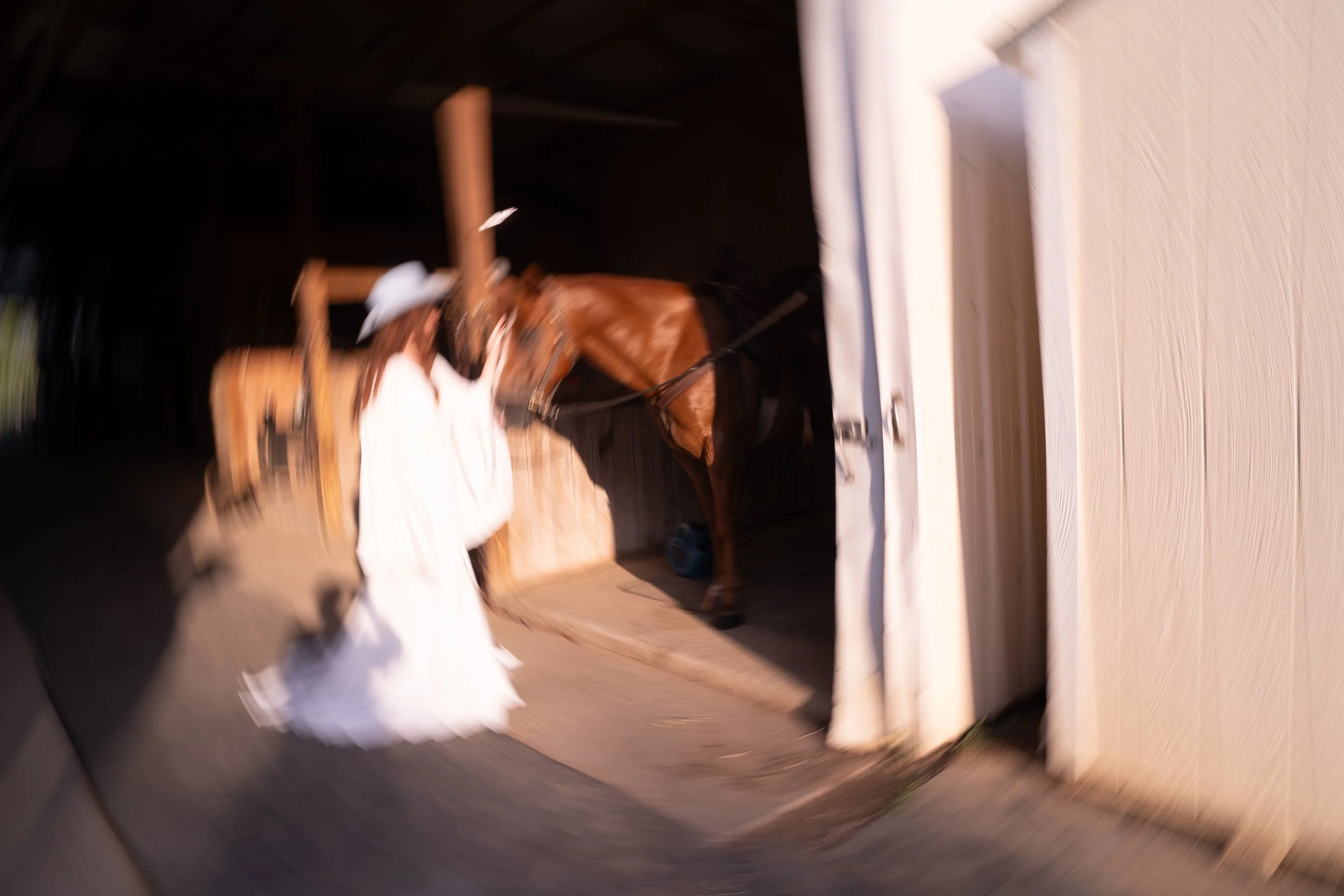 A woman in a white dress and large white hat standing in front of a brown horse in a stable.