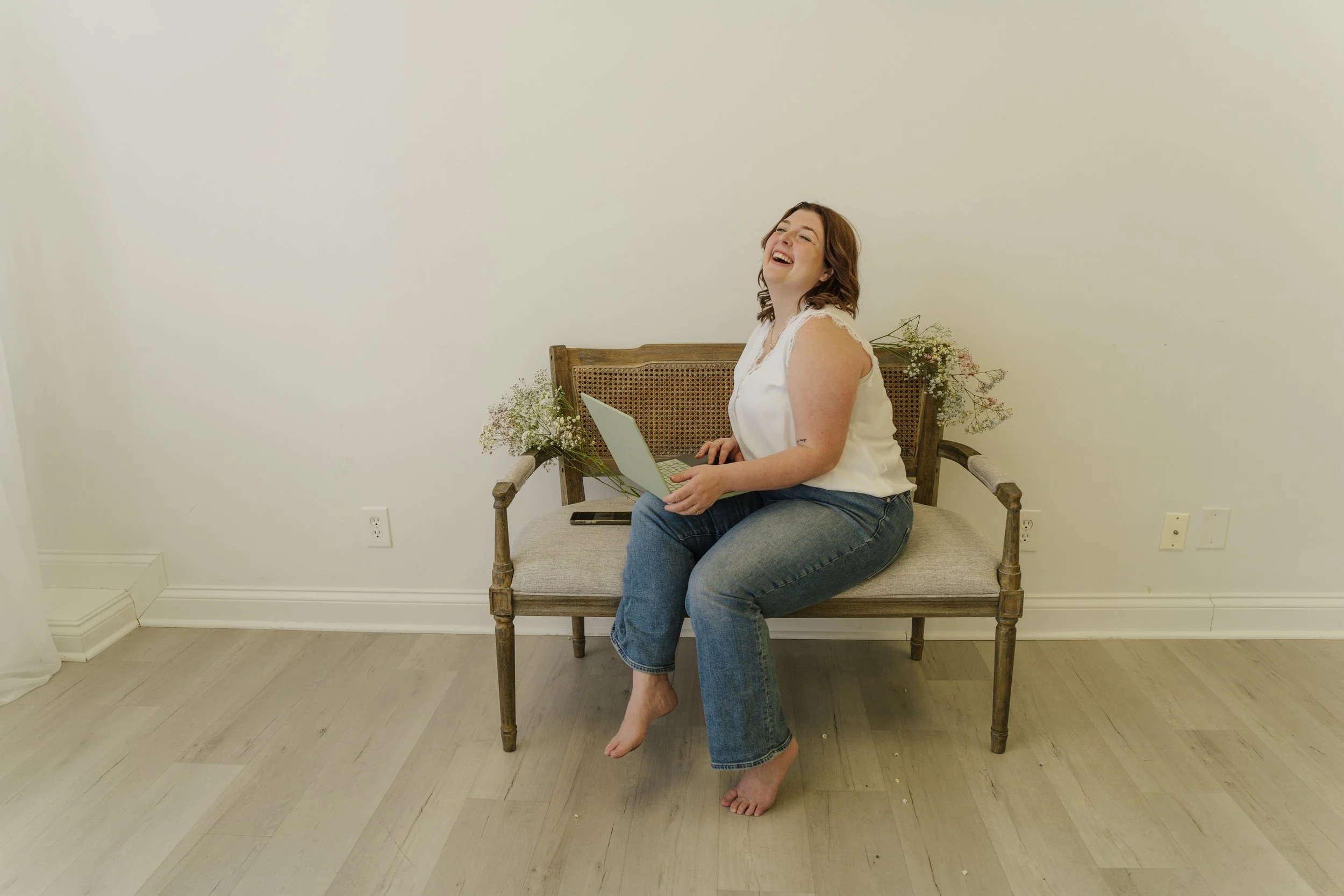 A woman with short reddish-brown hair, wearing a sleeveless white top and blue jeans, sitting on a wooden bench with her feet off the ground. She is holding a laptop and laughing. There are small floral arrangements on either side of the bench and a plain white wall behind her.