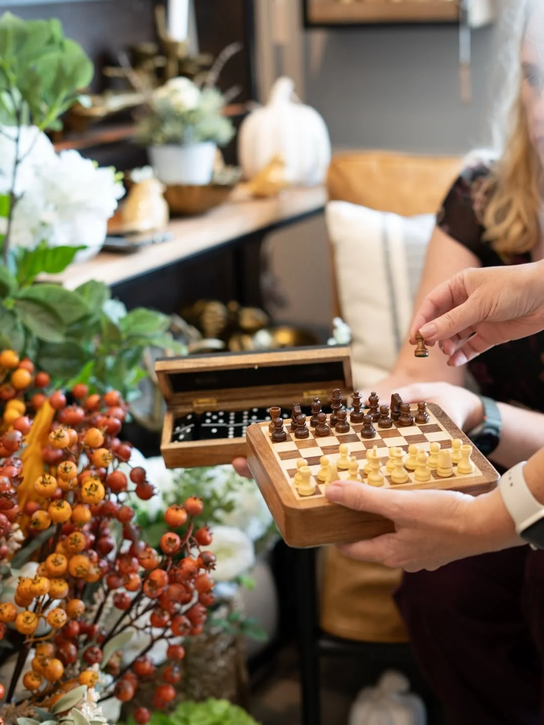 Person playing chess with a wooden chess set on a table surrounded by plants and flowers indoors.