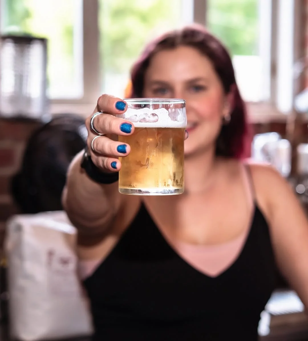 Woman with blue painted nails holding a glass of beer indoors, with a blurred background and natural light coming through windows.