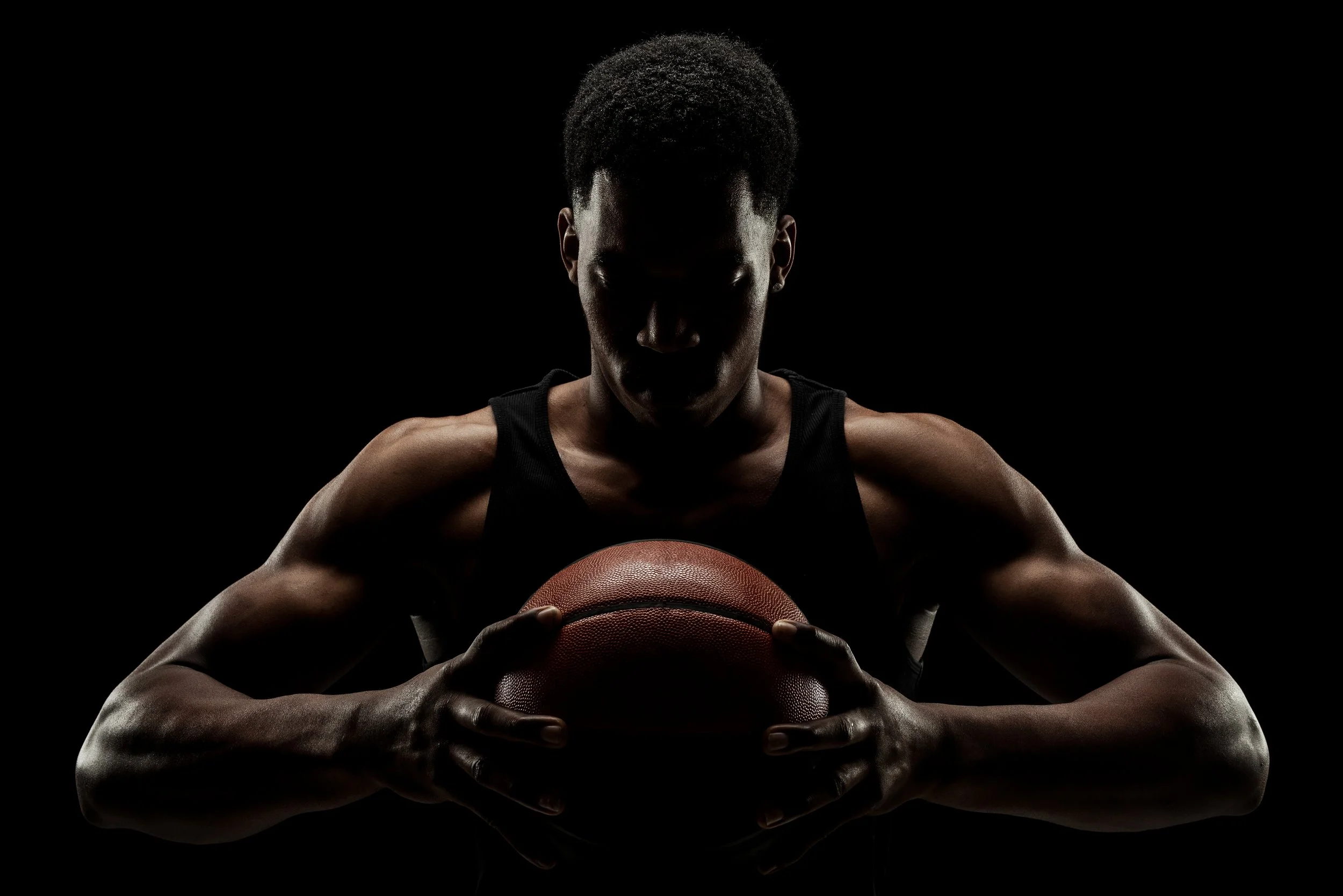 A dark silhouette of a young male athlete holding a basketball, with arms flexed and wearing a sleeveless sports jersey, against a black background.
