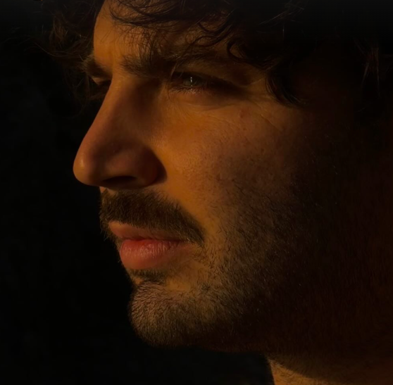Side profile of a man's face with dark, curly hair and facial hair, looking into the distance.
