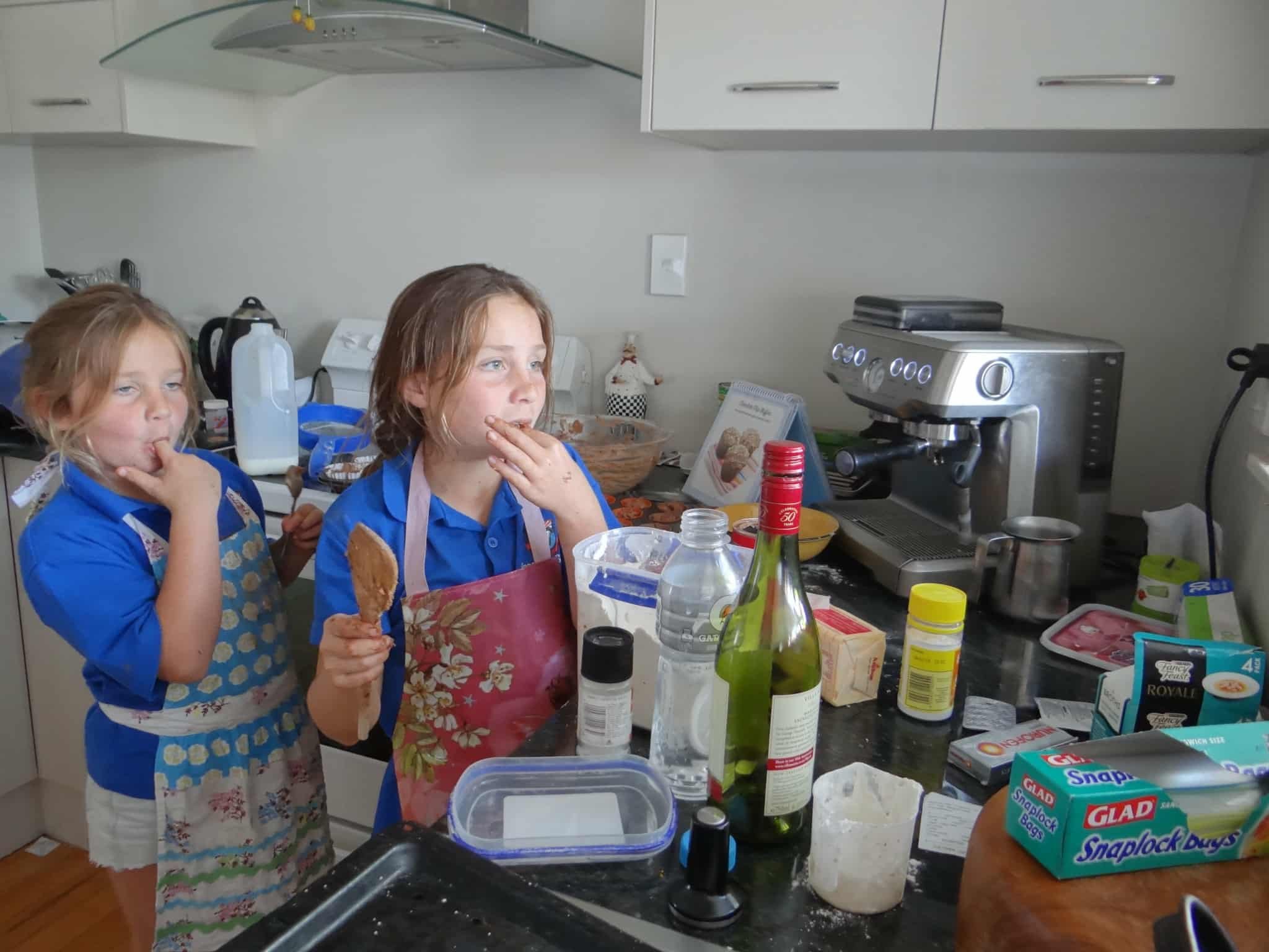 Two young children in blue shirts and aprons tasting food while preparing cookies in a kitchen with a coffee machine and baking supplies on the counter.