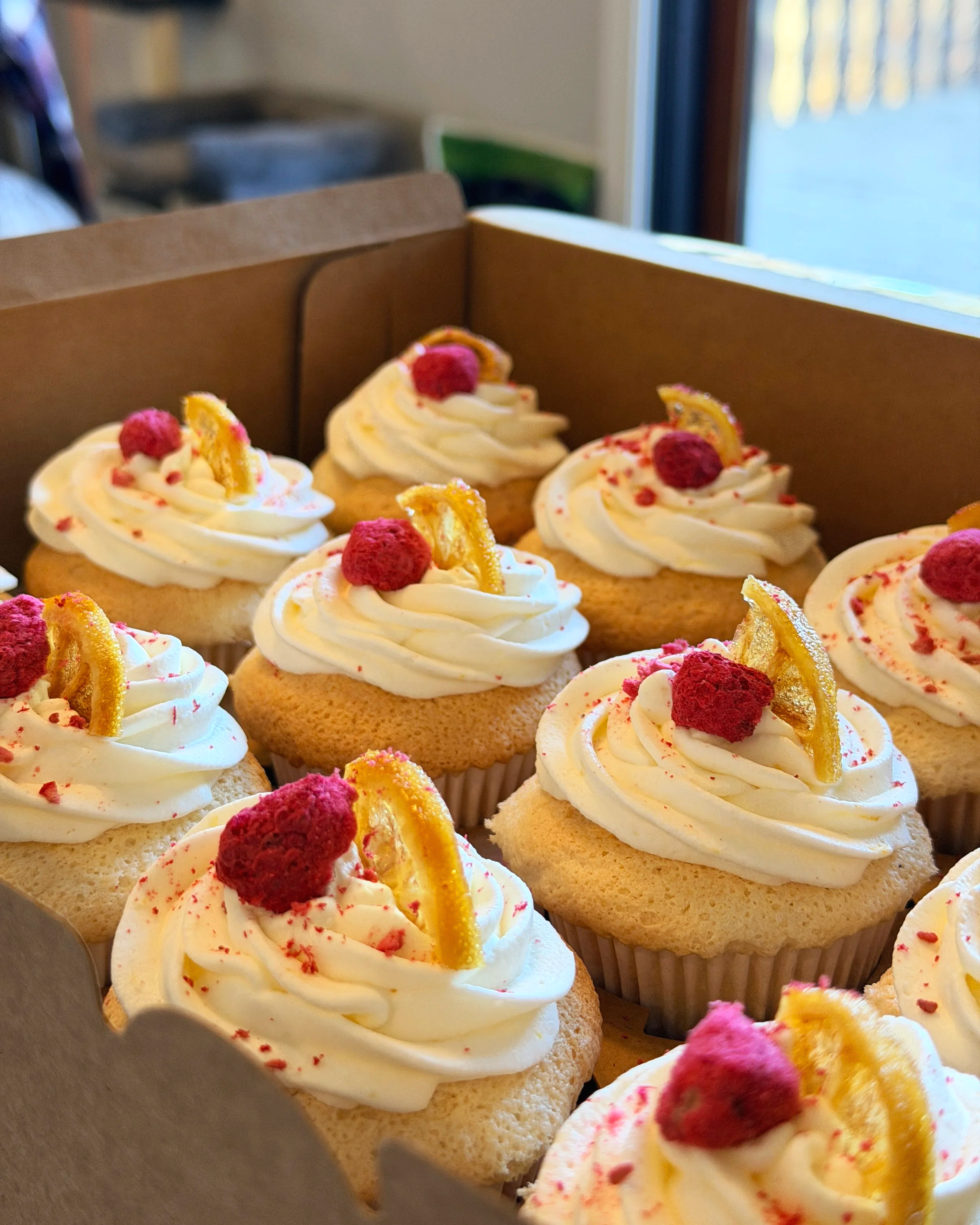 A box of angel food cupcakes with white frosting, topped with freeze dried raspberries and lemon slices. Made as a custom order bake good for sunflour bakehouse in Red Deer.