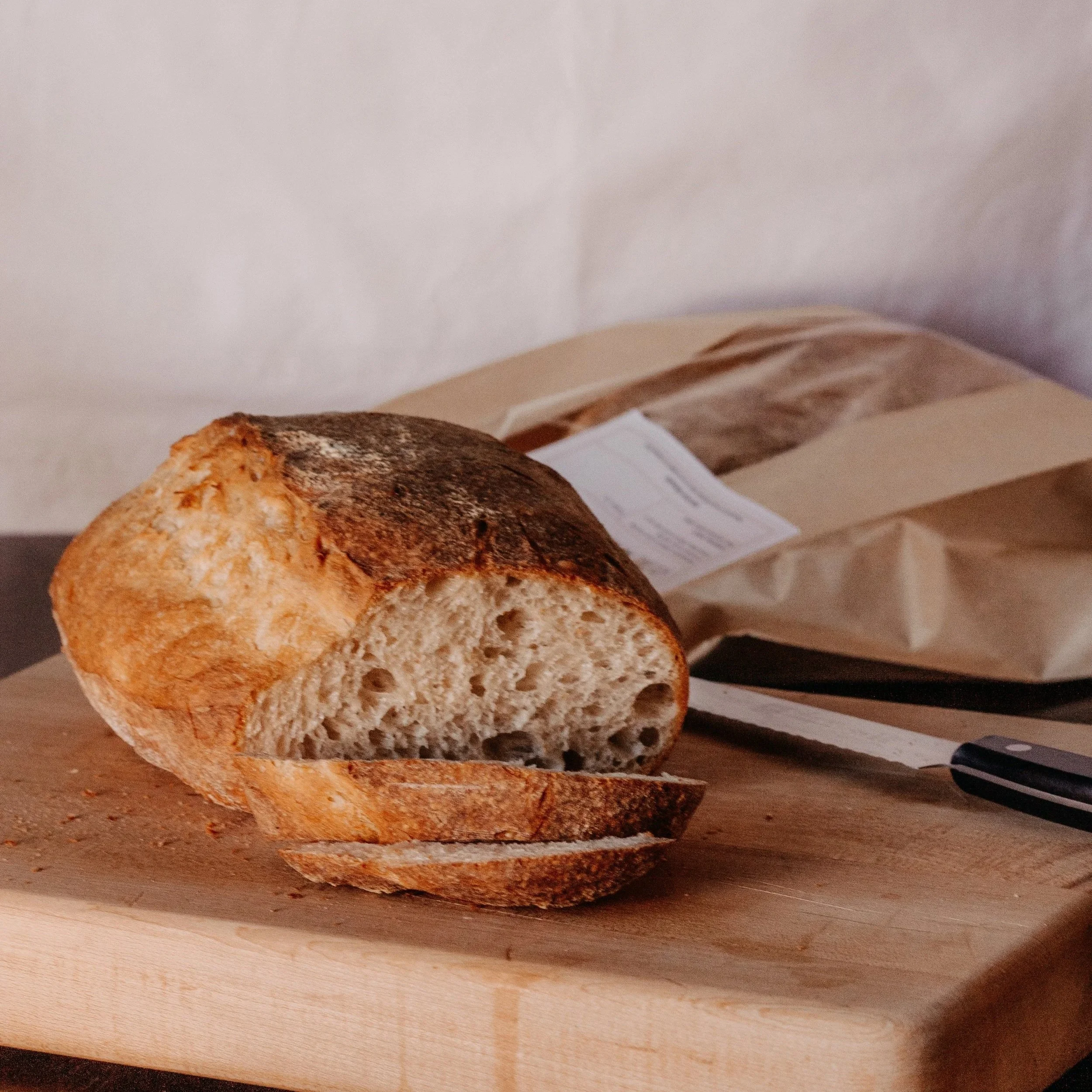 Sliced loaf of crusty artisan sourdough bread on a wooden cutting board with a bread knife and a paper bag in the background that was made in a home-based bakery near red deer.