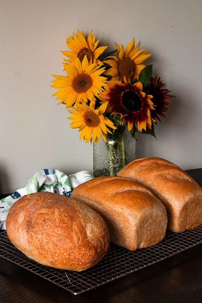 Three loaves of freshly baked bread resting on a wire cooling rack, with a bouquet of vibrant sunflowers in a vase in the background.