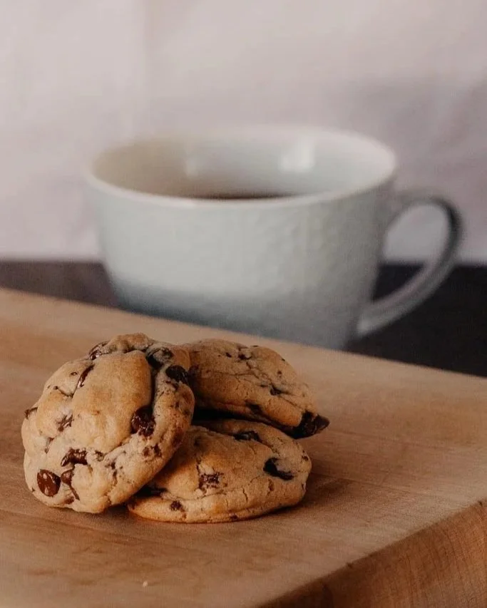 Close-up of three homemade, quality chocolate chip cookies on a wooden surface with a white mug in the background.