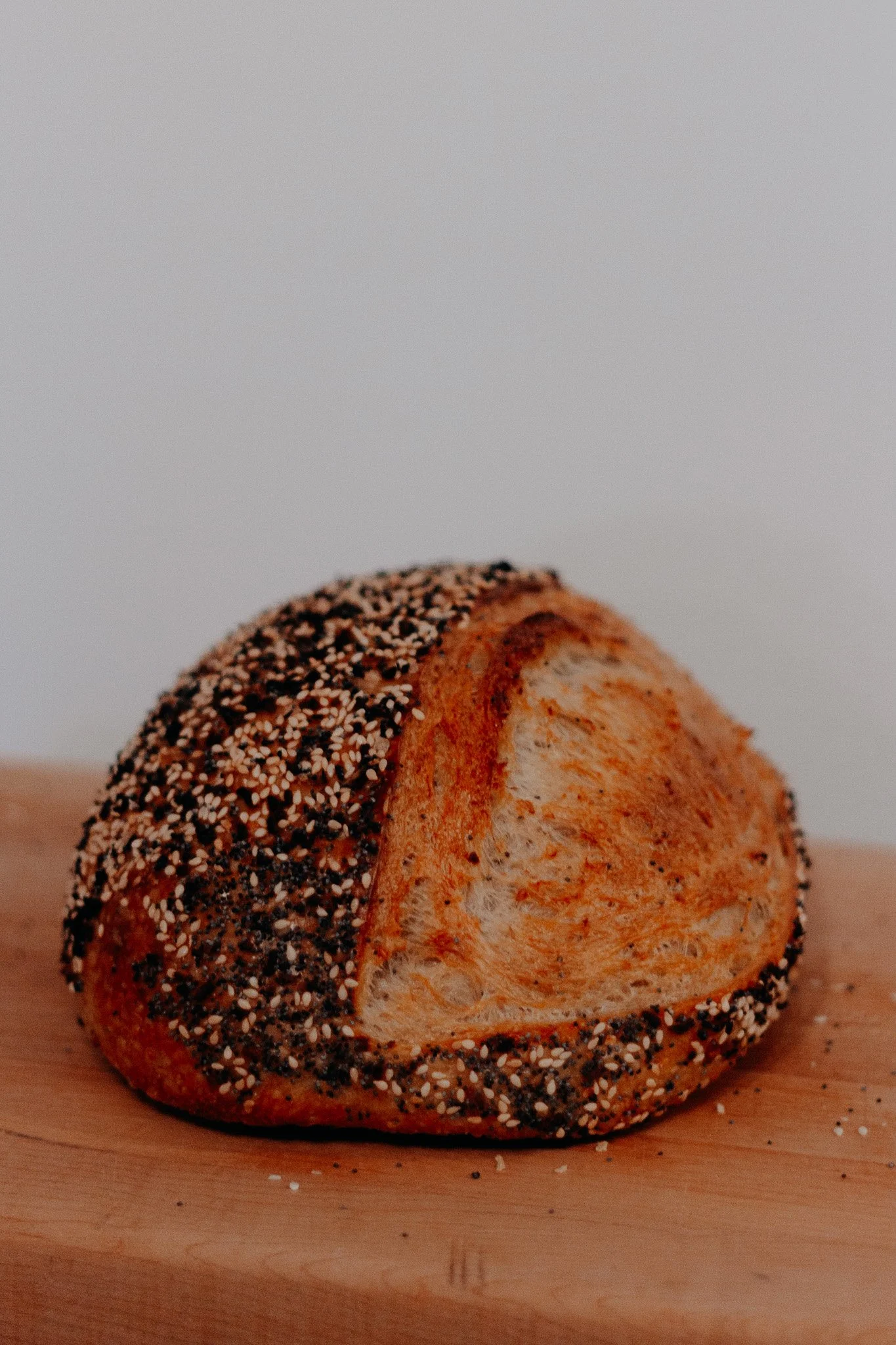 A round artisian sourdough bread loaf, with a crusted top, sprinkled with sesame and other seeds to mimic a everthing bagel seasoning, resting on a wooden surface in the sunflour bakehouse, bakery based in the Red Deer County area