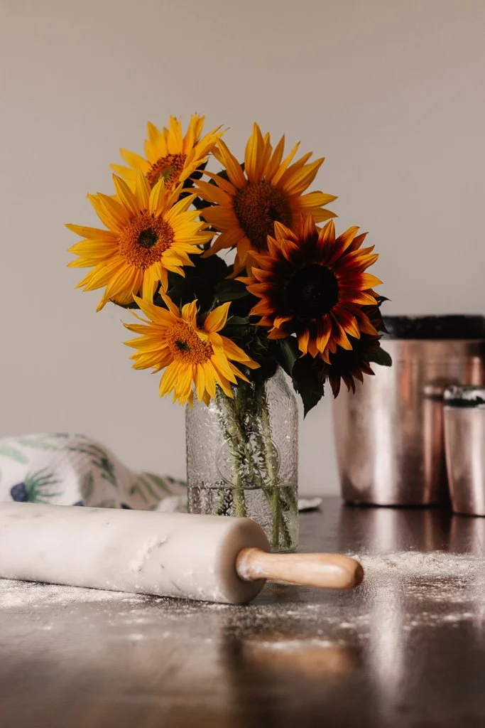 A glass vase with a bouquet of sunflowers on a kitchen counter, with a rolling pin and metal containers nearby.