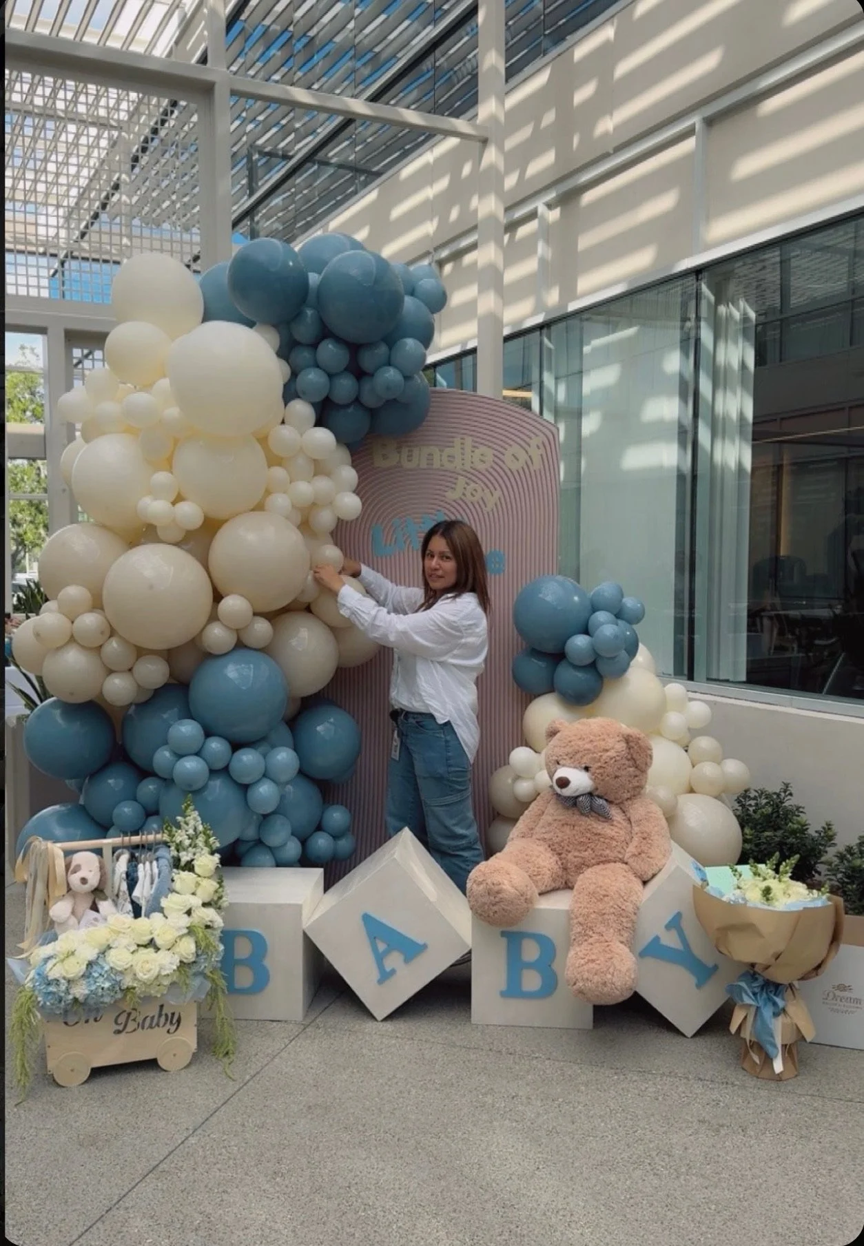 A woman standing next to a baby-themed decoration setup with blue, white, and cream-colored balloons, large toy blocks spelling 'BABY,' a teddy bear, and flower arrangements, inside a building with glass walls and a metal ceiling.