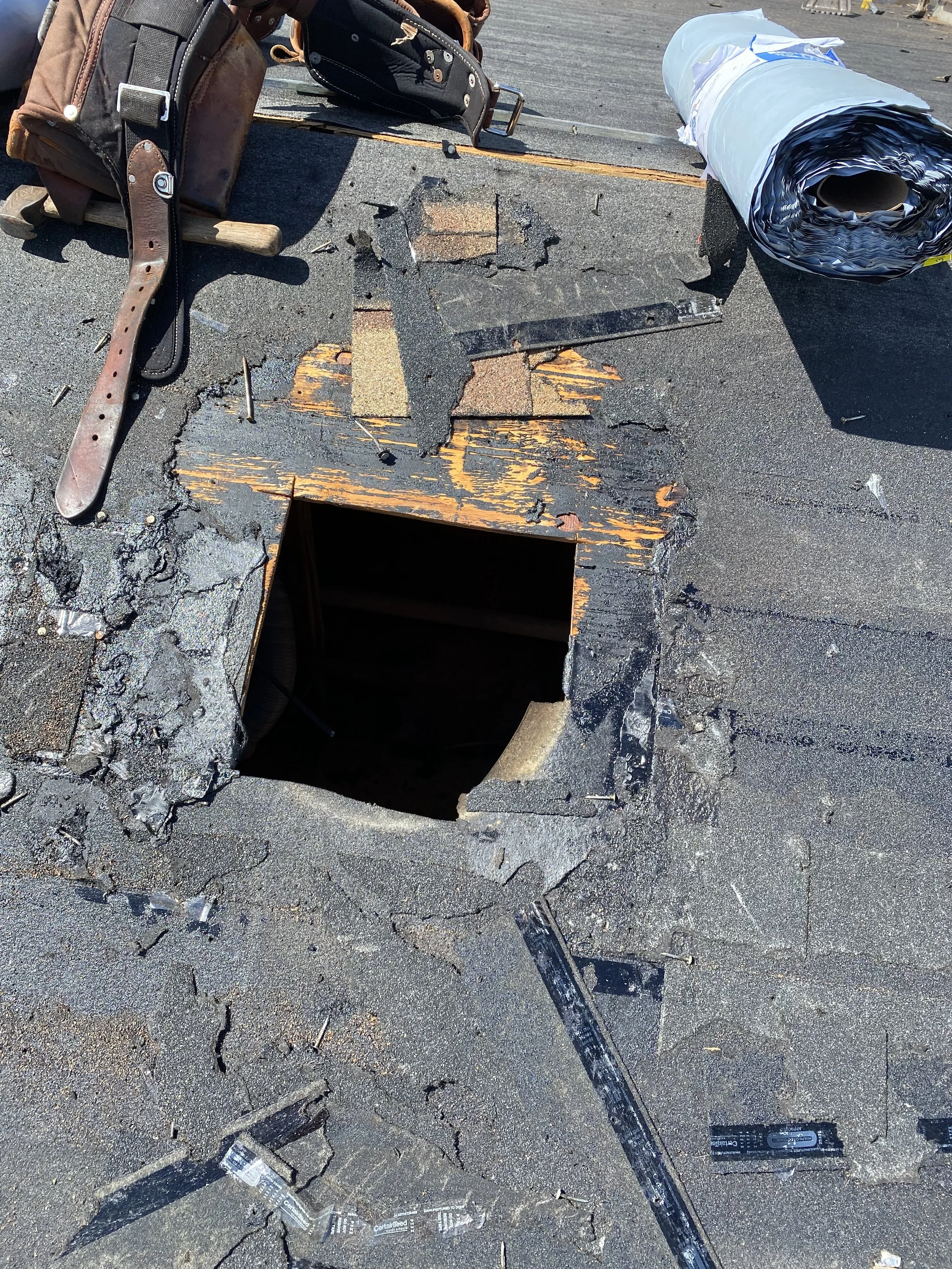 Roof with a large hole and scattered debris, tools, and rolled-up roofing material nearby.