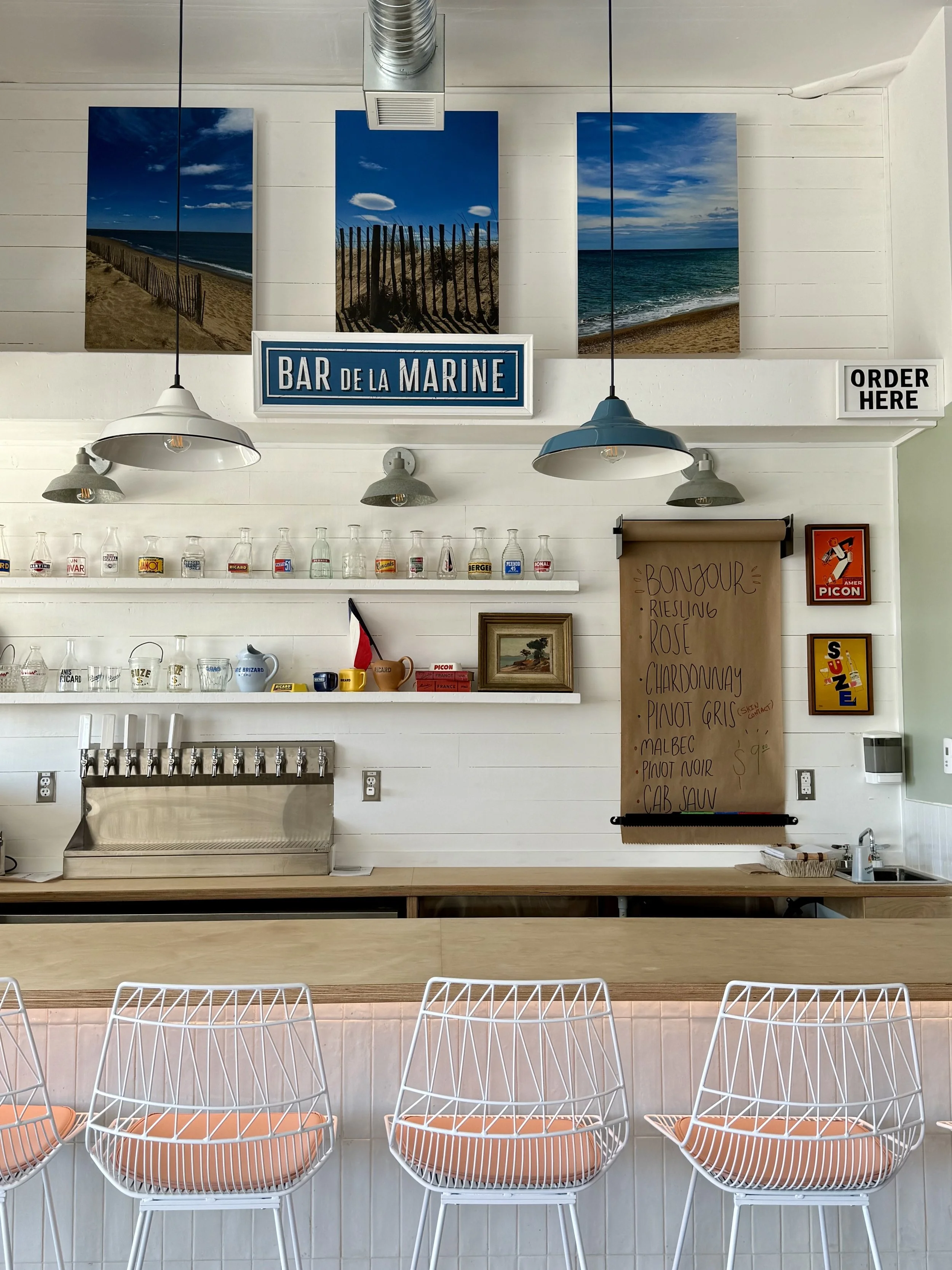Interior of a beach-themed bar with white walls, artwork of the beach, hanging lamps, shelves with glass bottles, and a menu board listing wines, with white bar stools with orange cushions.