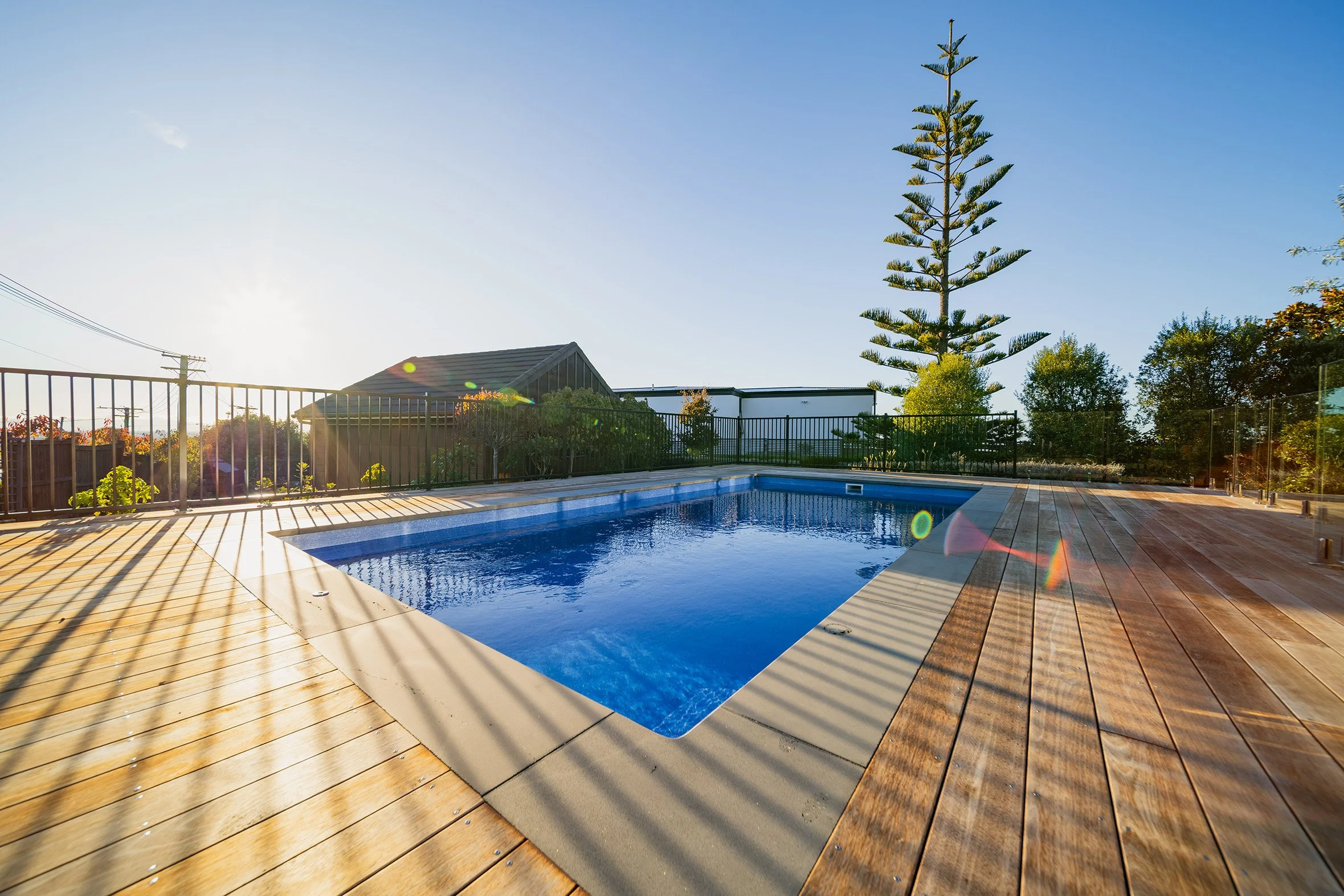 An outdoor swimming pool with a wooden deck, surrounded by a black fence, with trees and a building in the background under a clear blue sky.