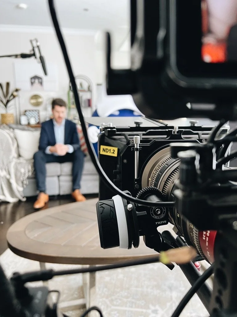 A behind the scenes photo of a camera on a tripod with an Angenieux lens filming a man in a suit sitting on a sofa in a living room.