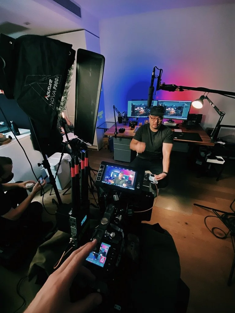 A video production setup with a camera filming a man in a room with dual computer monitors and studio lighting, while Blake Castle operates a RED video camera in the background.