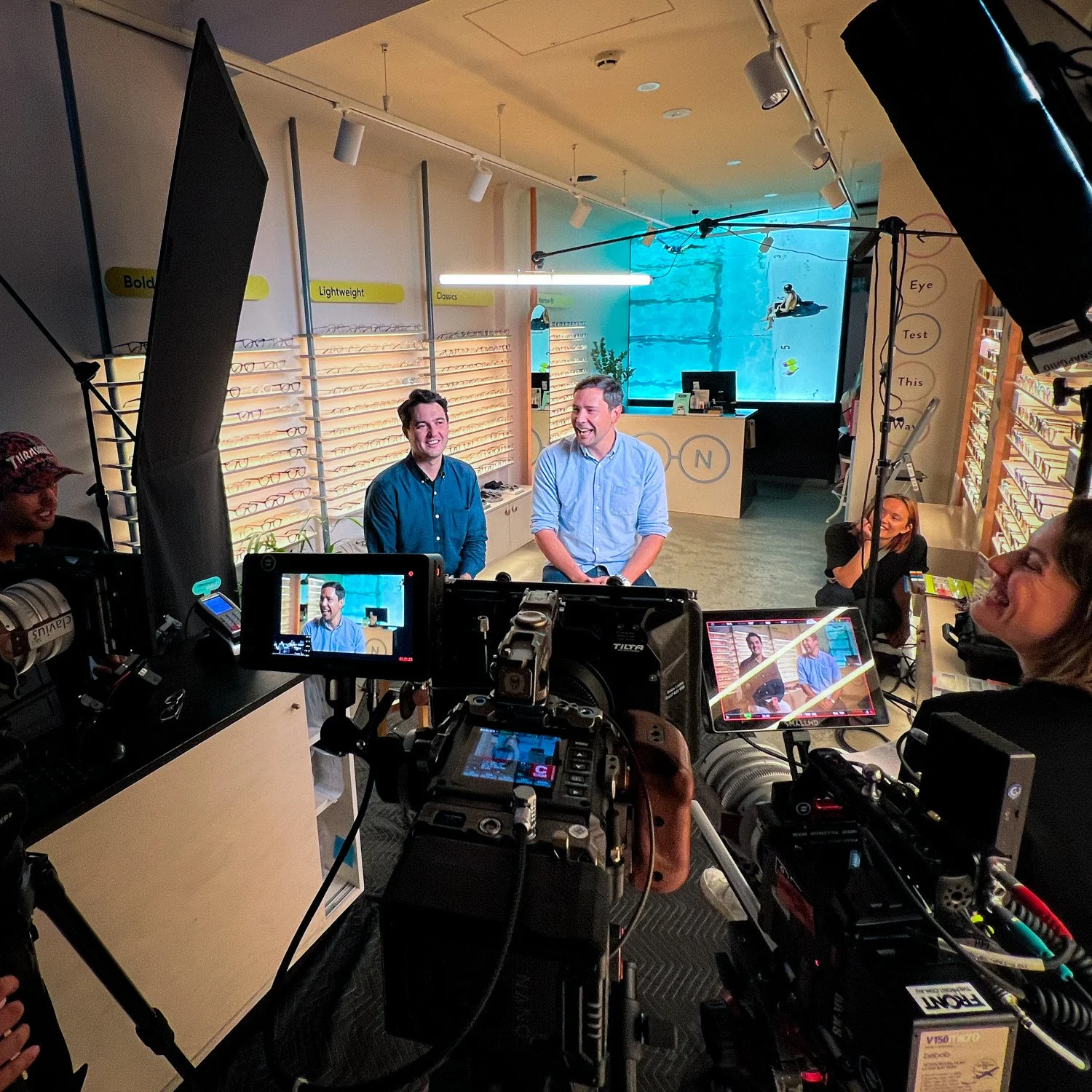 Two men sitting and speaking as interviewees in front of cameras and lighting equipment inside the Bailey Nelson eyeglass store, with display shelves of eyeglasses in the background.