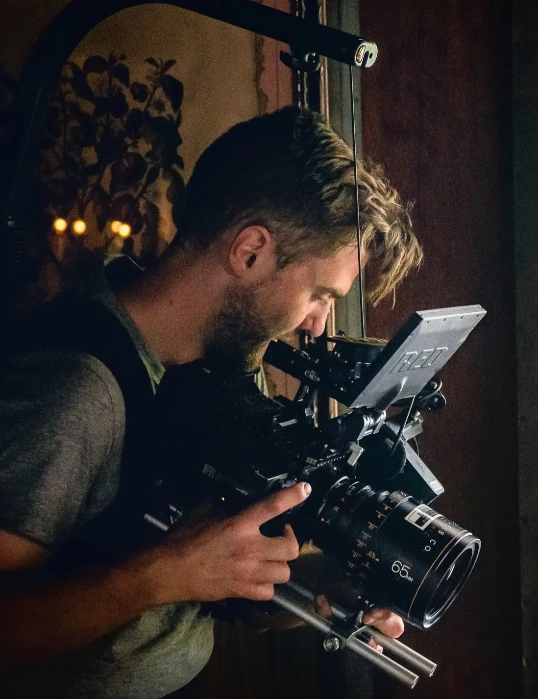 Blake Castle operating a RED cinema camera with an anamorphic lens attached to an easyrig while looking at the monitor, indoors with warm lighting.