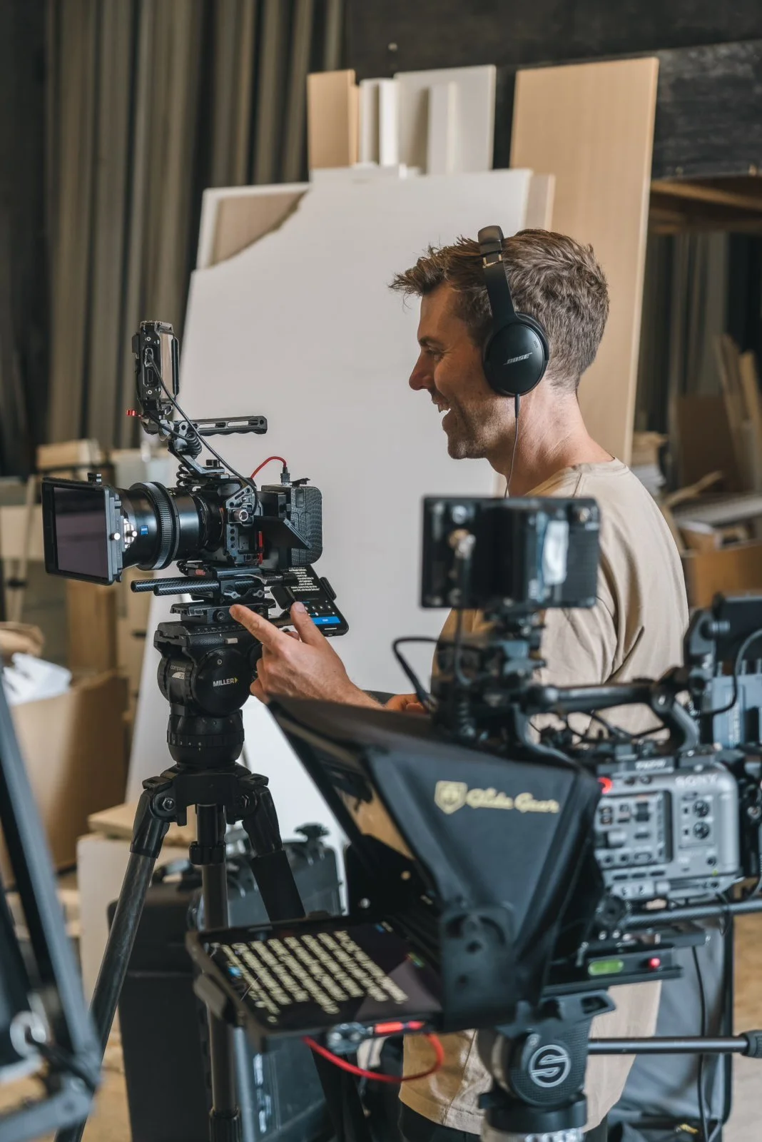 Blake castle wearing headphones operates a professional video camera on a tripod, as well as a second camera with a teleprompter in a workshop setting with wooden and white panels in the background.