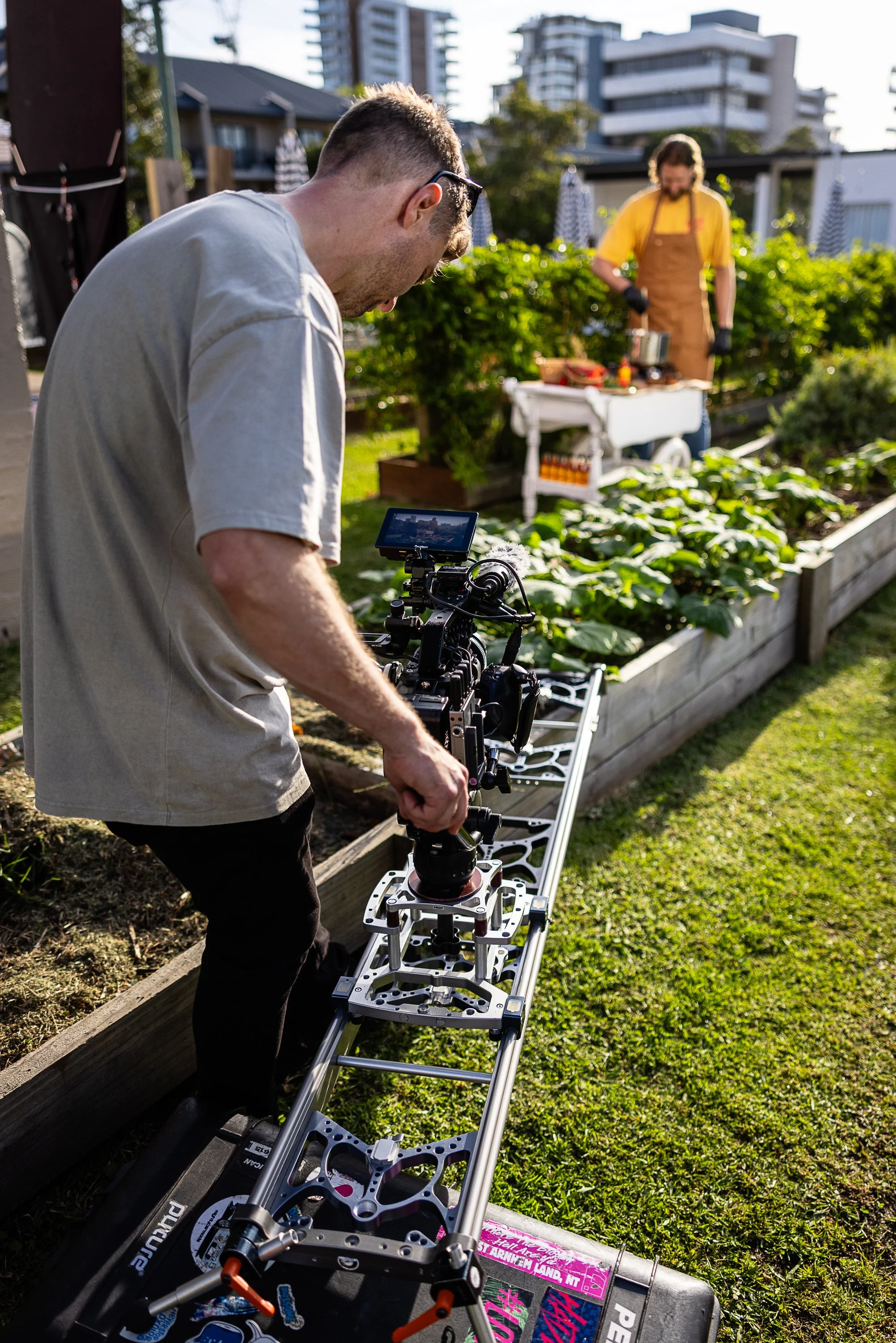 Blake Castle operating a cinema camera on a slider in a garden, with a man in a yellow shirt and apron cooking an overly large pot of hot sauce in the garden.