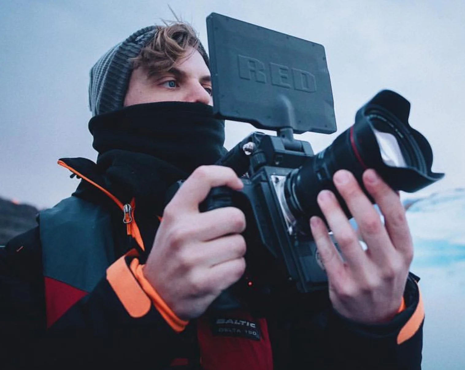 Blake Castle operating a RED cinema camera filming a documentary in Greenland with icebergs in the background, looking at the monitor of the camera wearing a large snow jacket and beanie.