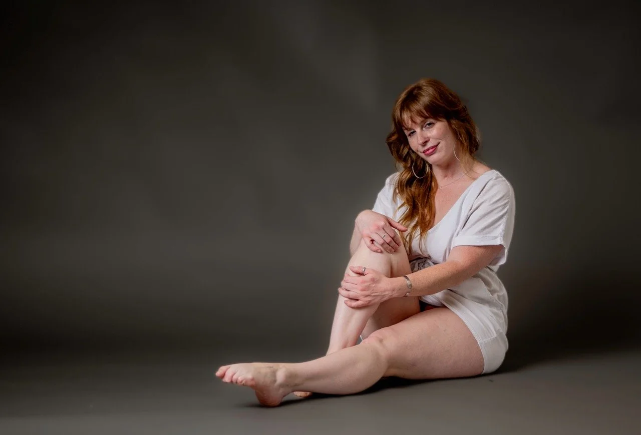 A woman with long red hair sitting on the floor against a dark background, wearing a white outfit, smiling, and holding her leg.