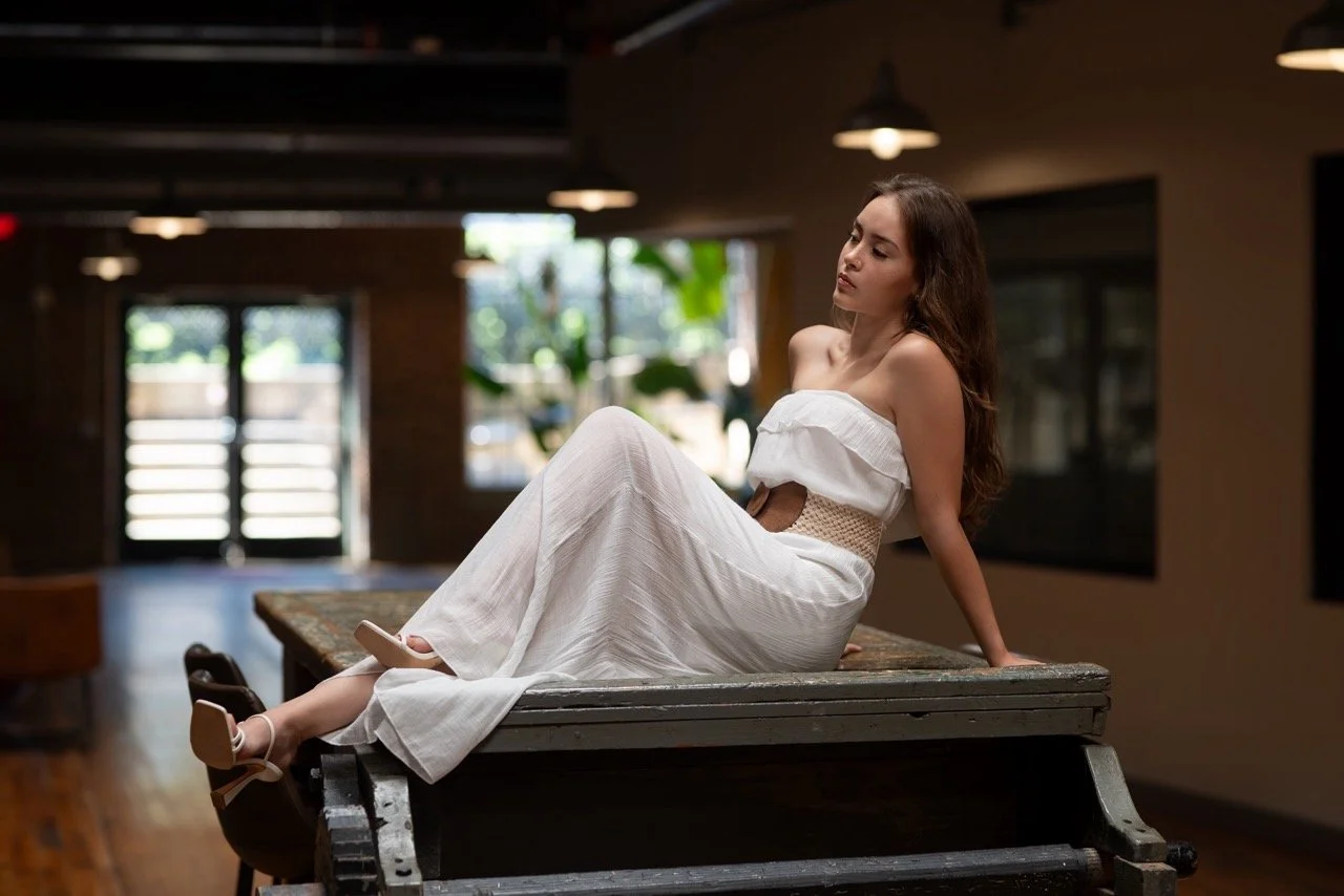 Woman in white dress sitting on a table in a room with large windows and warm lighting.