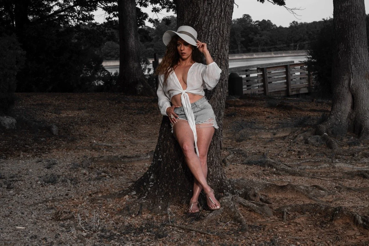 A woman with long curly hair wearing a large white sun hat, a white tied-up blouse, and distressed denim shorts, is leaning against a tree in a park near a body of water at sunset or dusk.