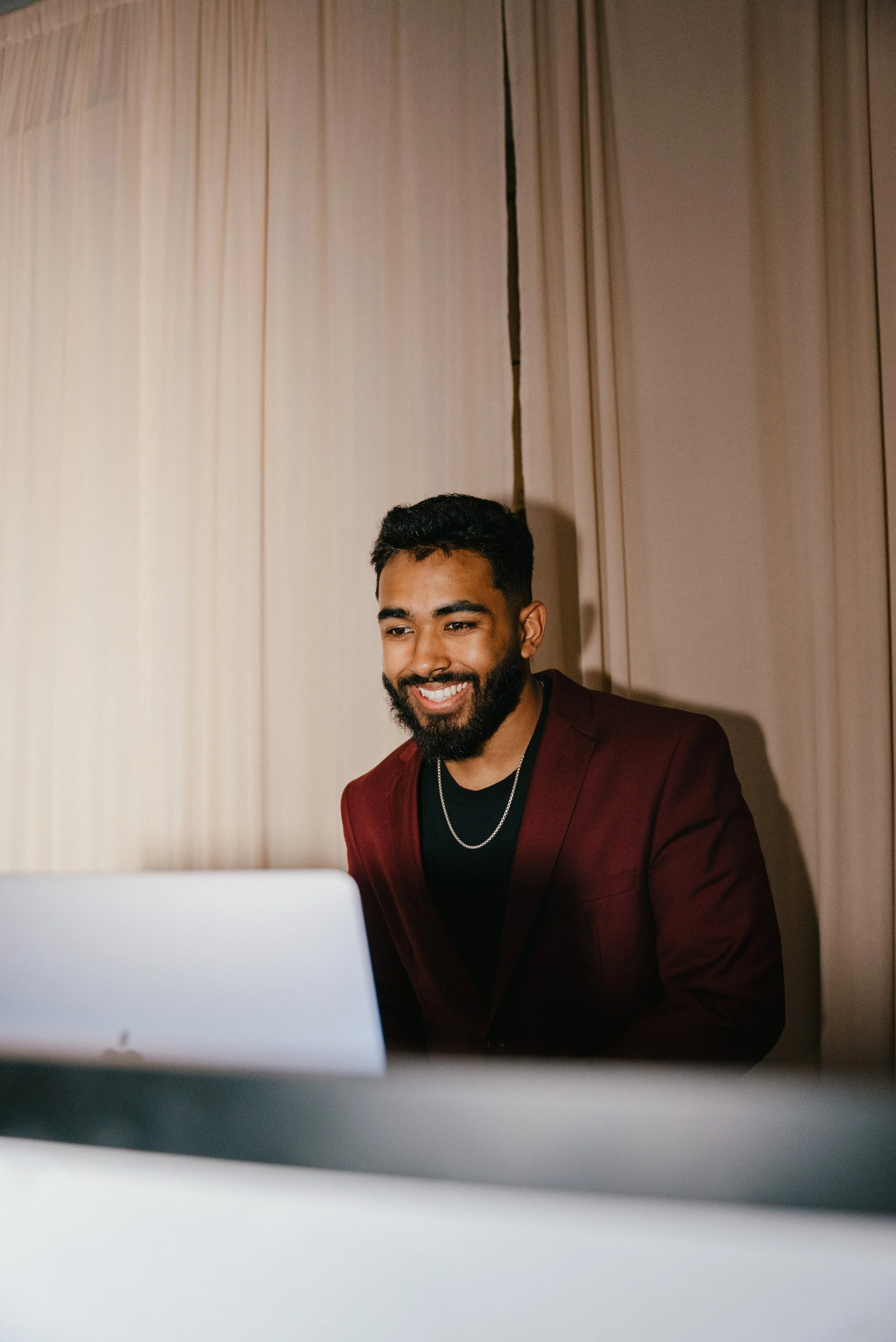 A smiling man with a beard and dark hair, wearing a maroon blazer, black shirt, and silver chain, looking at a laptop screen in a room with beige curtains.