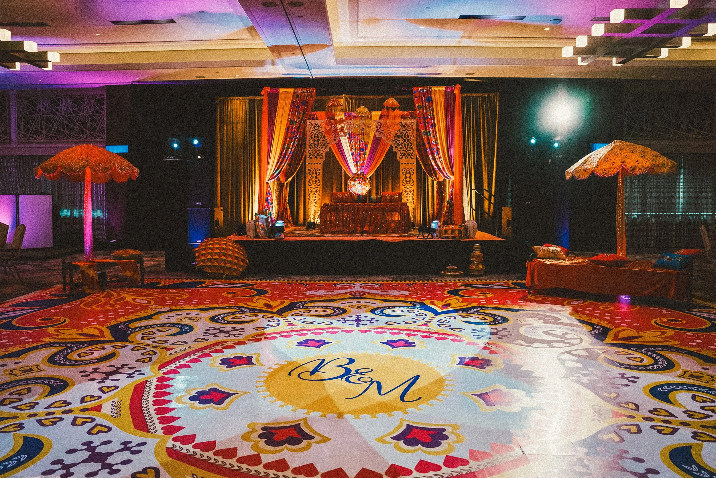 An elaborately decorated event stage with a colorful, ornate carpet in the foreground featuring a monogram with the initials 'B' and 'M'. The stage has vibrant, patterned drapes, and two large decorative umbrellas flanking it. The setting appears to be in a ballroom or banquet hall with subdued lighting and decorative lighting effects.