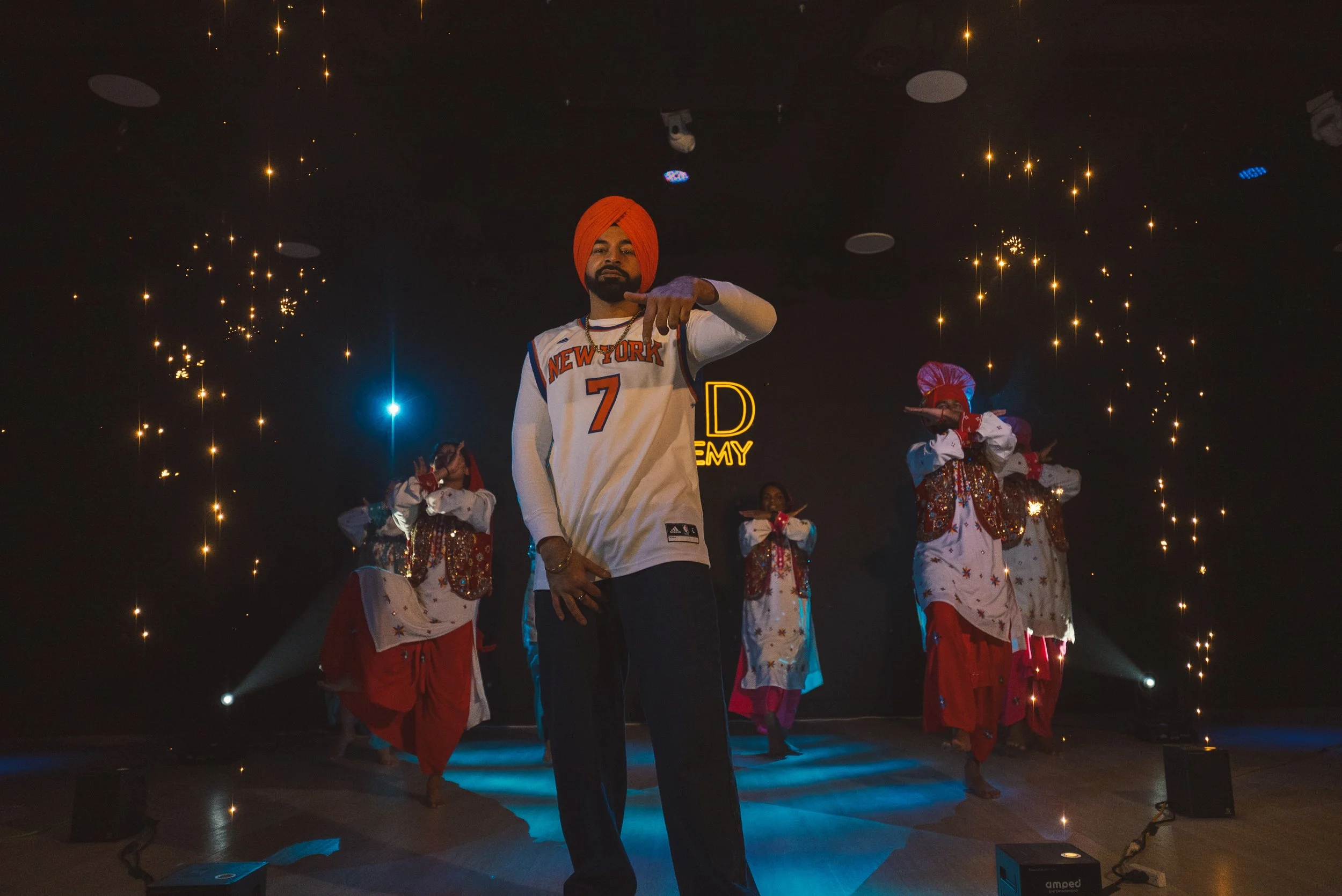 A man in a white basketball jersey with orange lettering and a red turban performing on stage with dancers dressed in traditional Indian attire behind him. The stage is decorated with bright lights and sparkles, with a neon sign in the background that reads 'DREAM.'