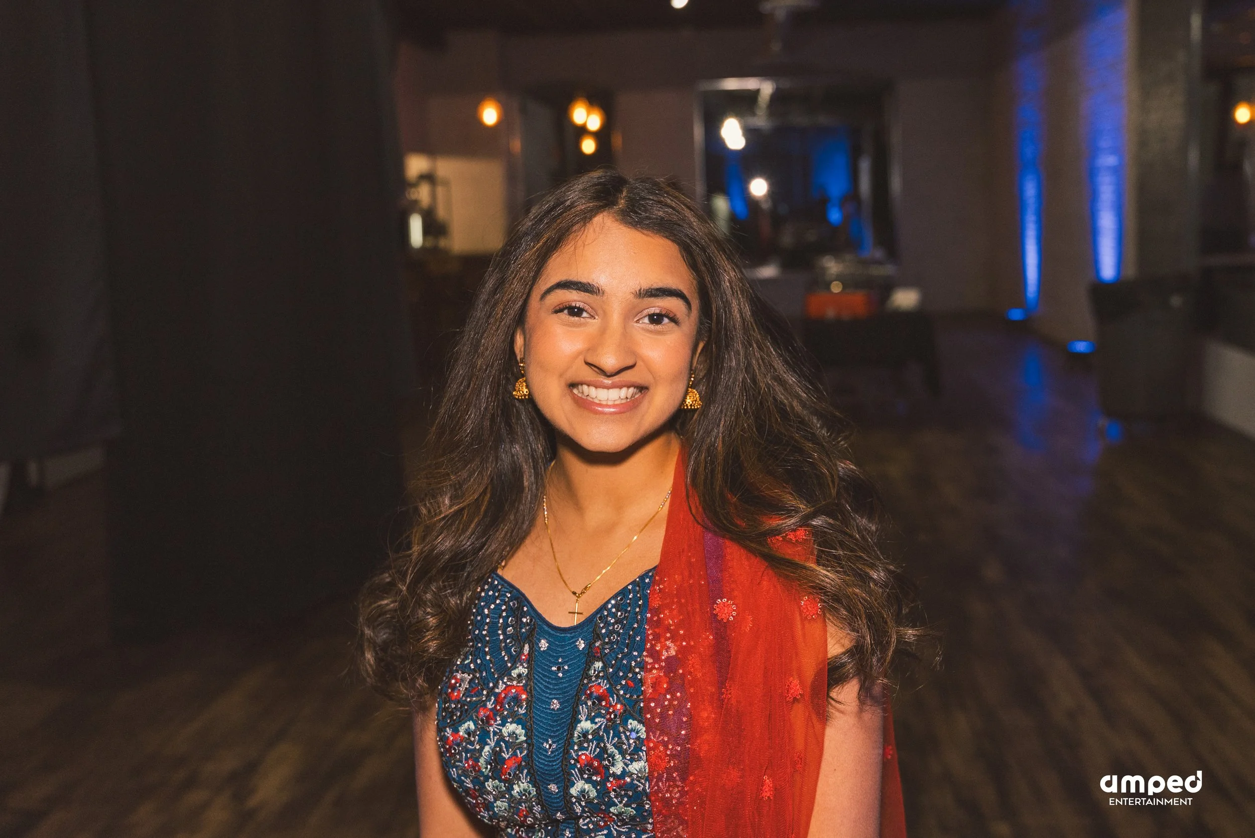 A smiling woman with long dark hair, wearing a colorful floral dress and gold jewelry, standing in a dimly lit indoor setting.