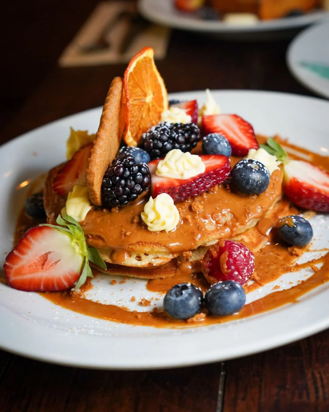 Plate of pancakes topped with assorted berries, sliced strawberries, whipped cream, caramel sauce, an orange slice, and cookies.