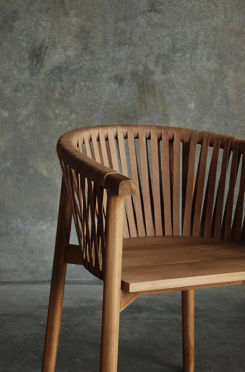 Wooden chair with a slatted backrest and armrest, against a textured concrete wall background.
