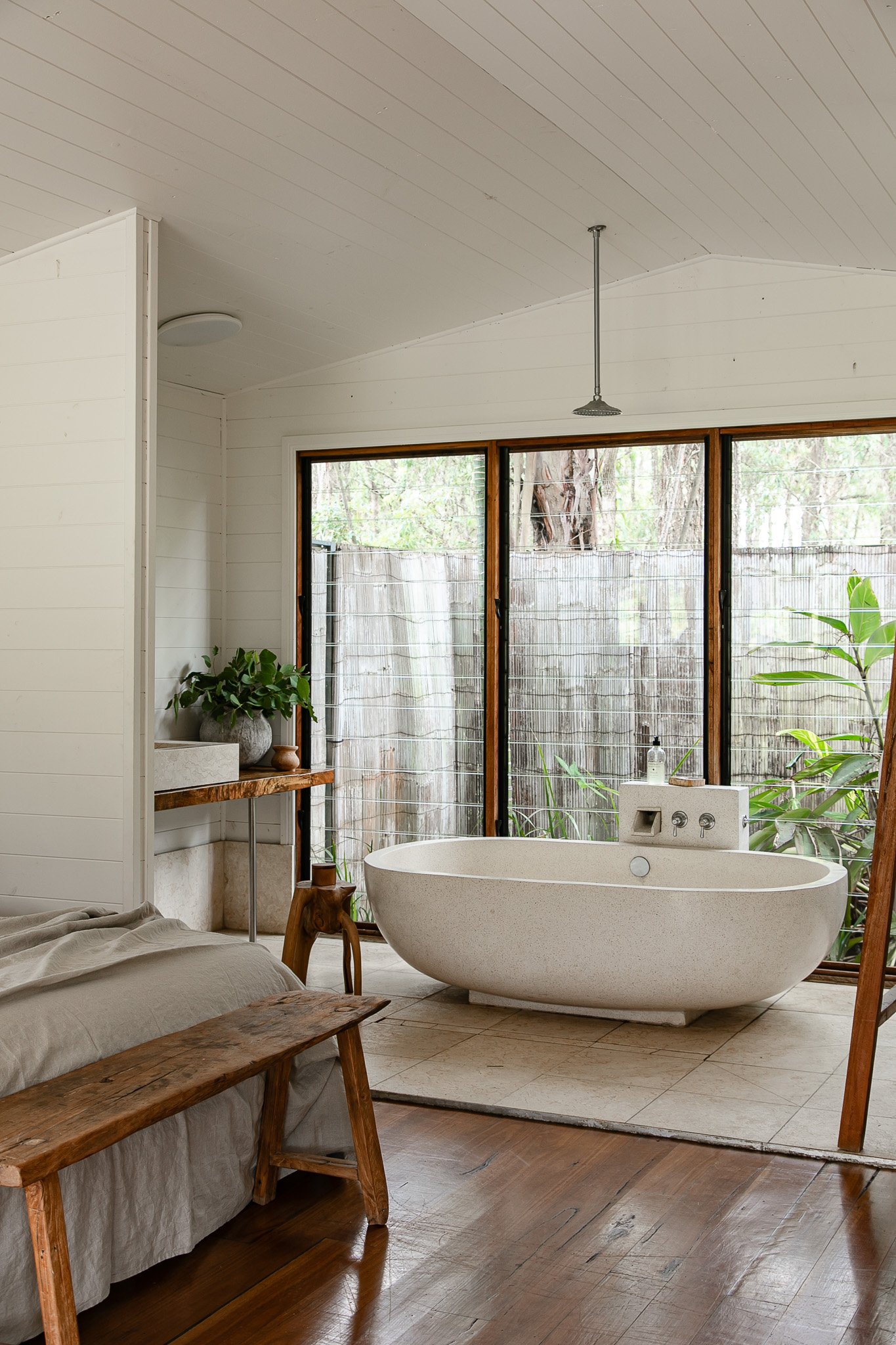 Interior view of a bathroom with a freestanding bathtub, large glass sliding doors, a wooden bench, and green plants outside.