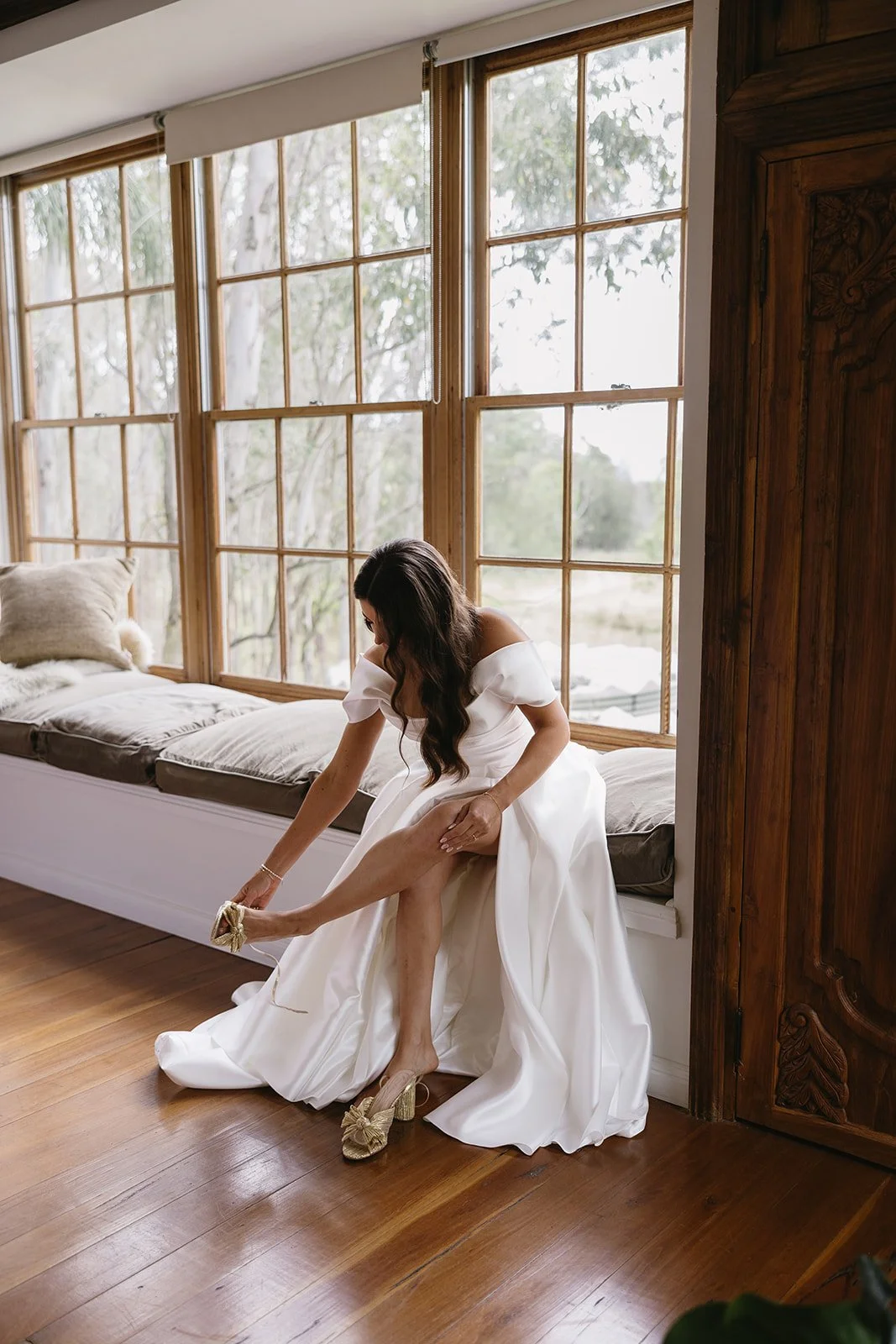 Bride in a white wedding dress sitting on a window seat, adjusting her gold high heel shoe.