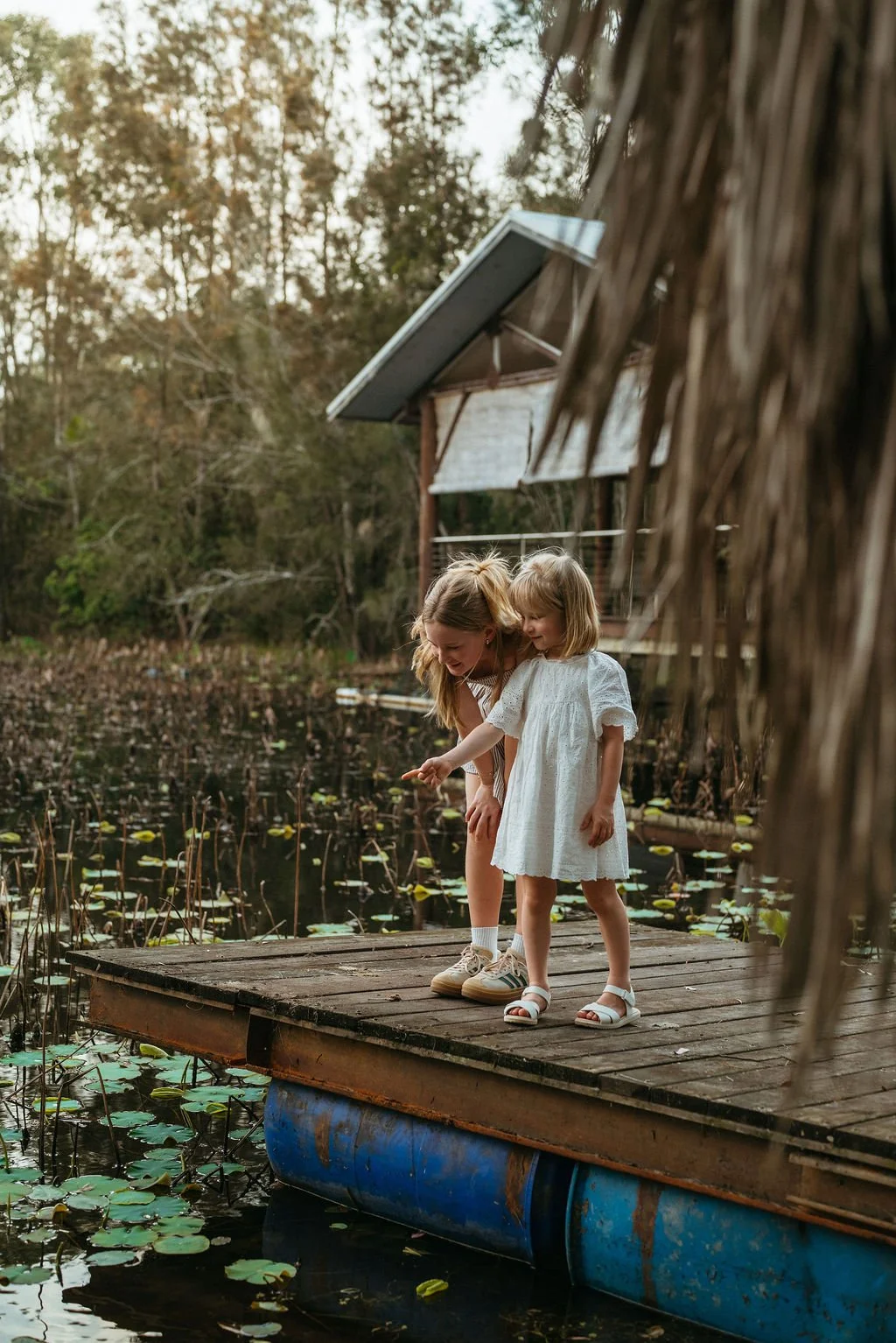 A woman and a young girl in white dresses standing on a dock by a pond, looking at the water and pointing. There are trees and a wooden house with a solar panel in the background at Leaves and Fishes